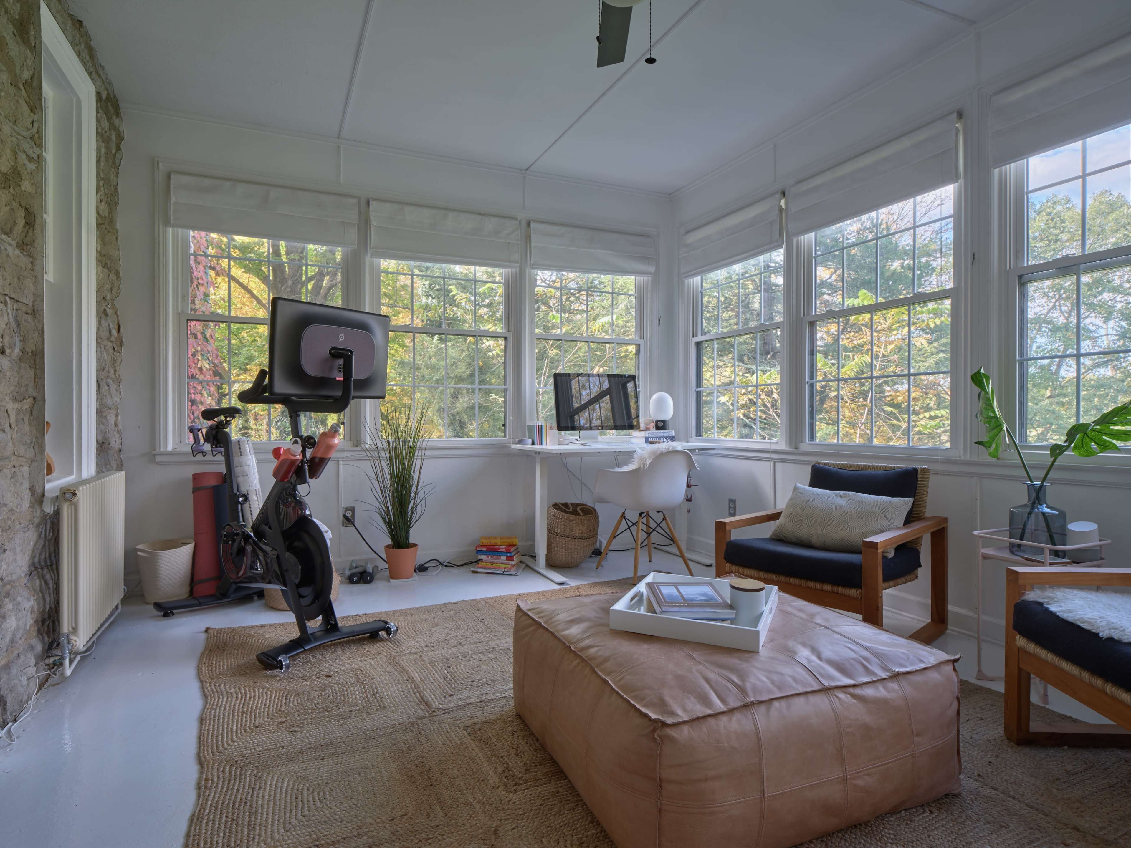 A sunlit room with large windows, featuring an exercise bike, a desk with a computer, a potted plant, and various seating options.