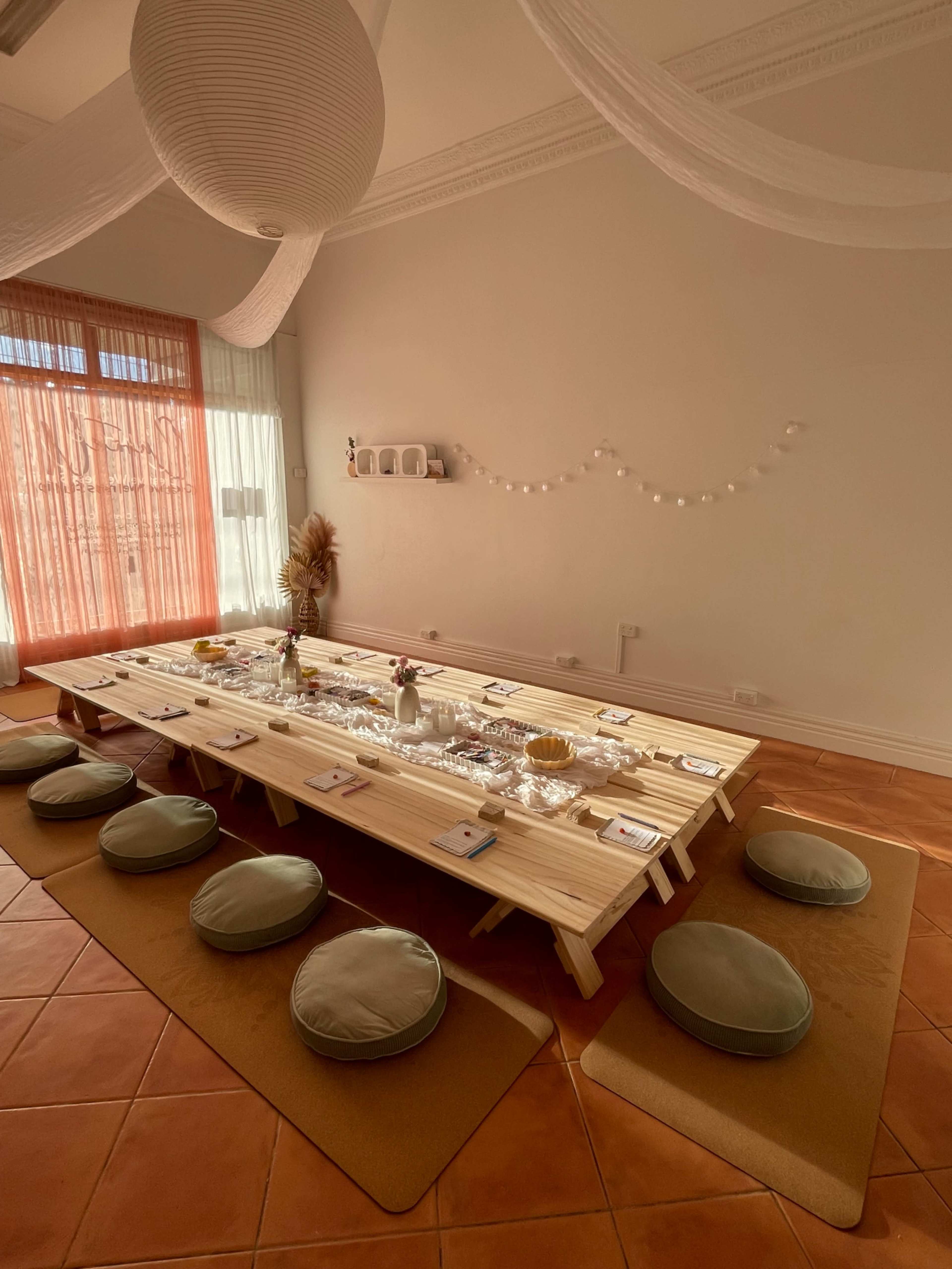 A long wooden table is set with place settings and decorative items in a bright, airy room with light-colored walls and terracotta tiled flooring.