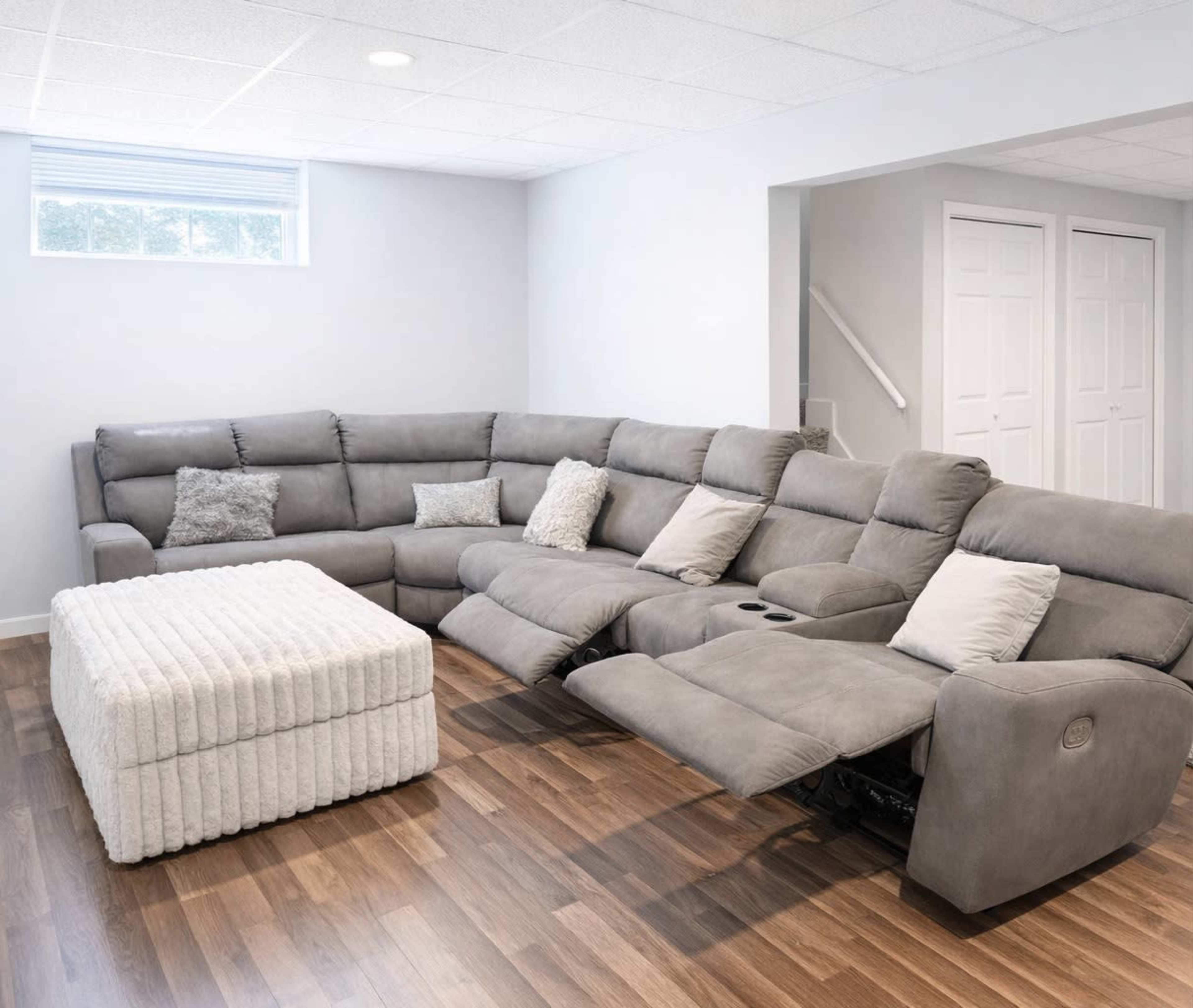 A spacious living room features a large gray sectional sofa with recliners and a textured white ottoman on a wooden floor.
