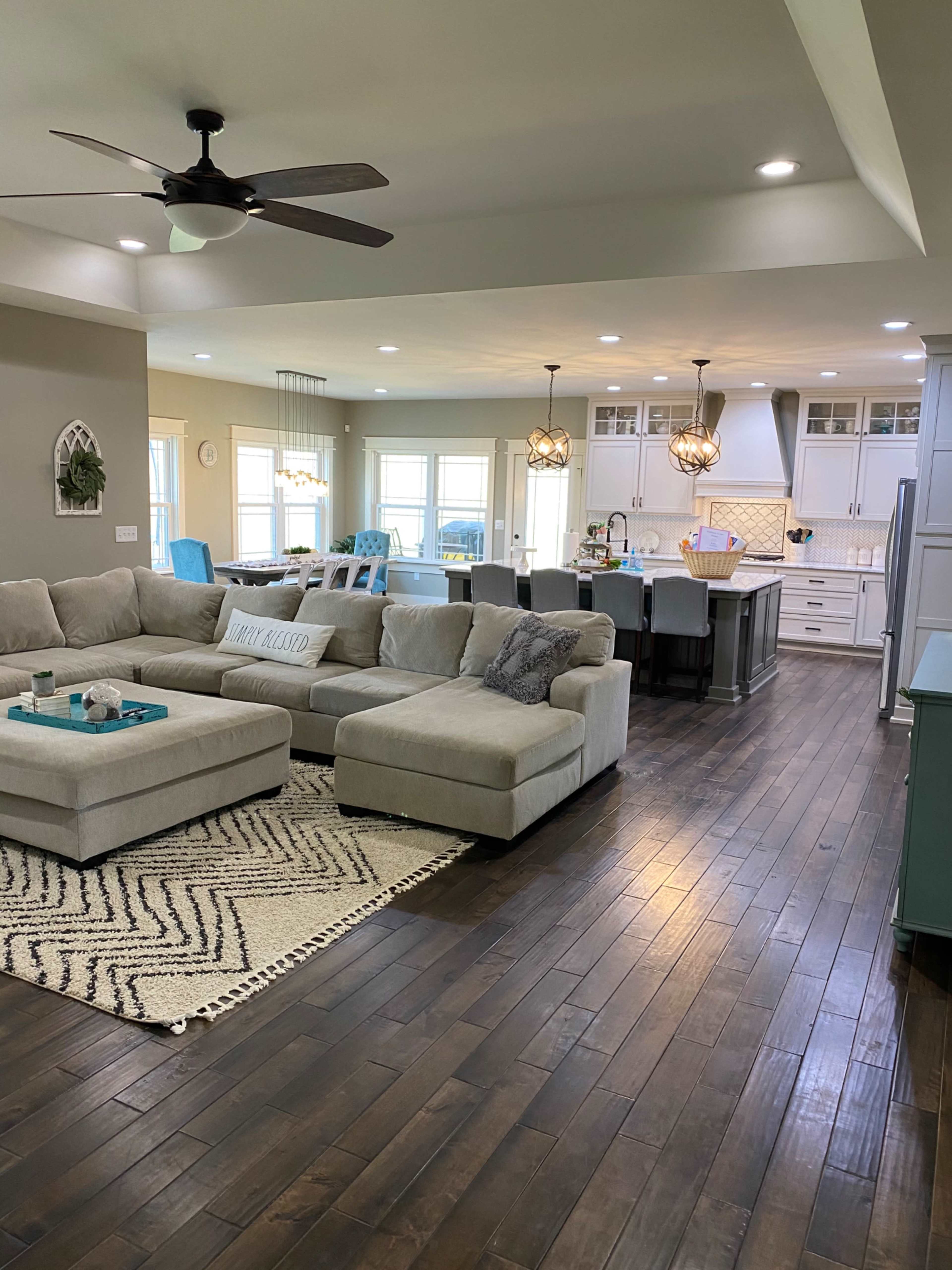 A modern living room with a large sectional sofa, a decorative rug, and an open kitchen in the background featuring a central island and pendant lighting.