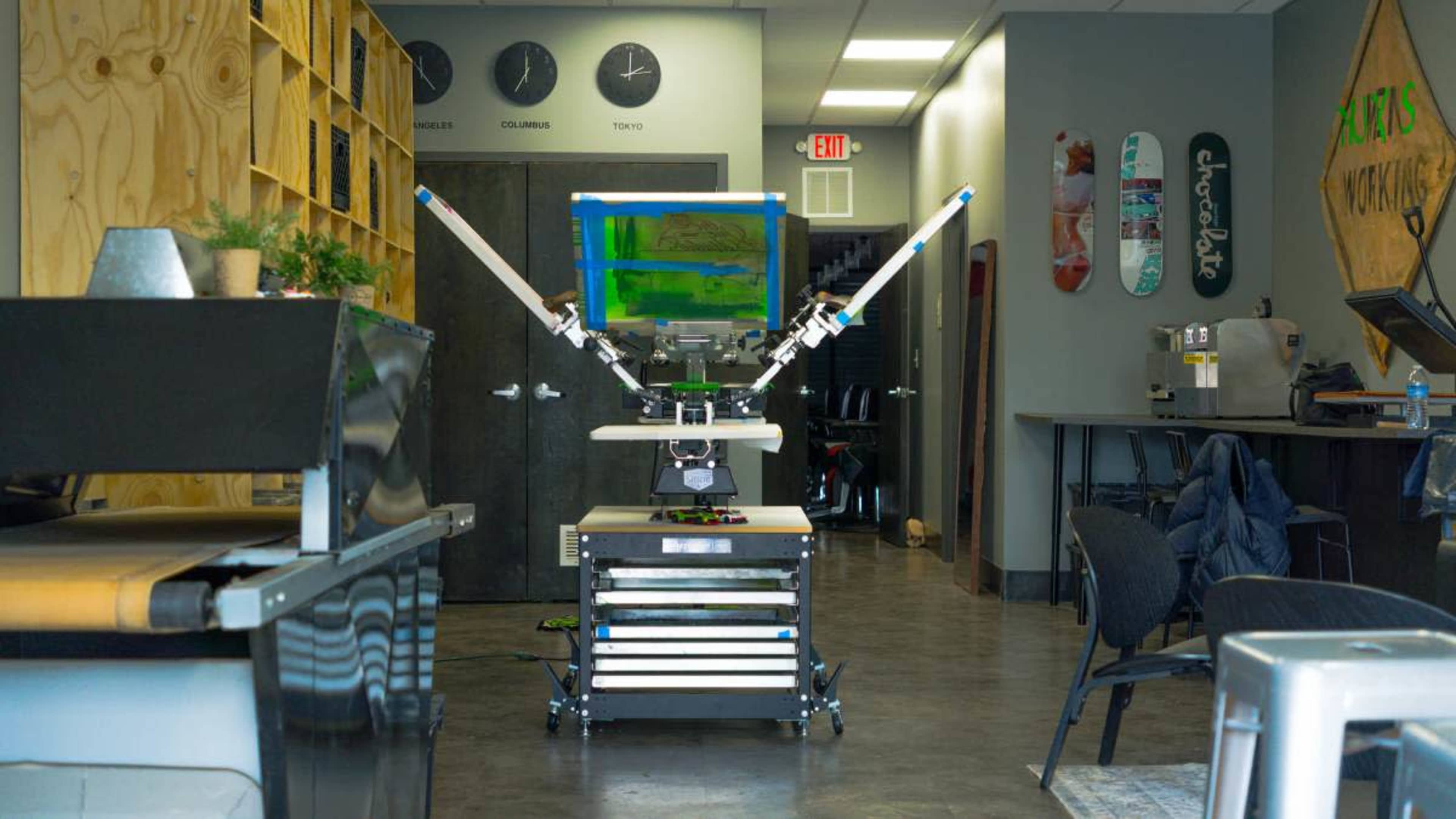 A screen printing machine is positioned in an industrial studio space, surrounded by various tools and furniture.
