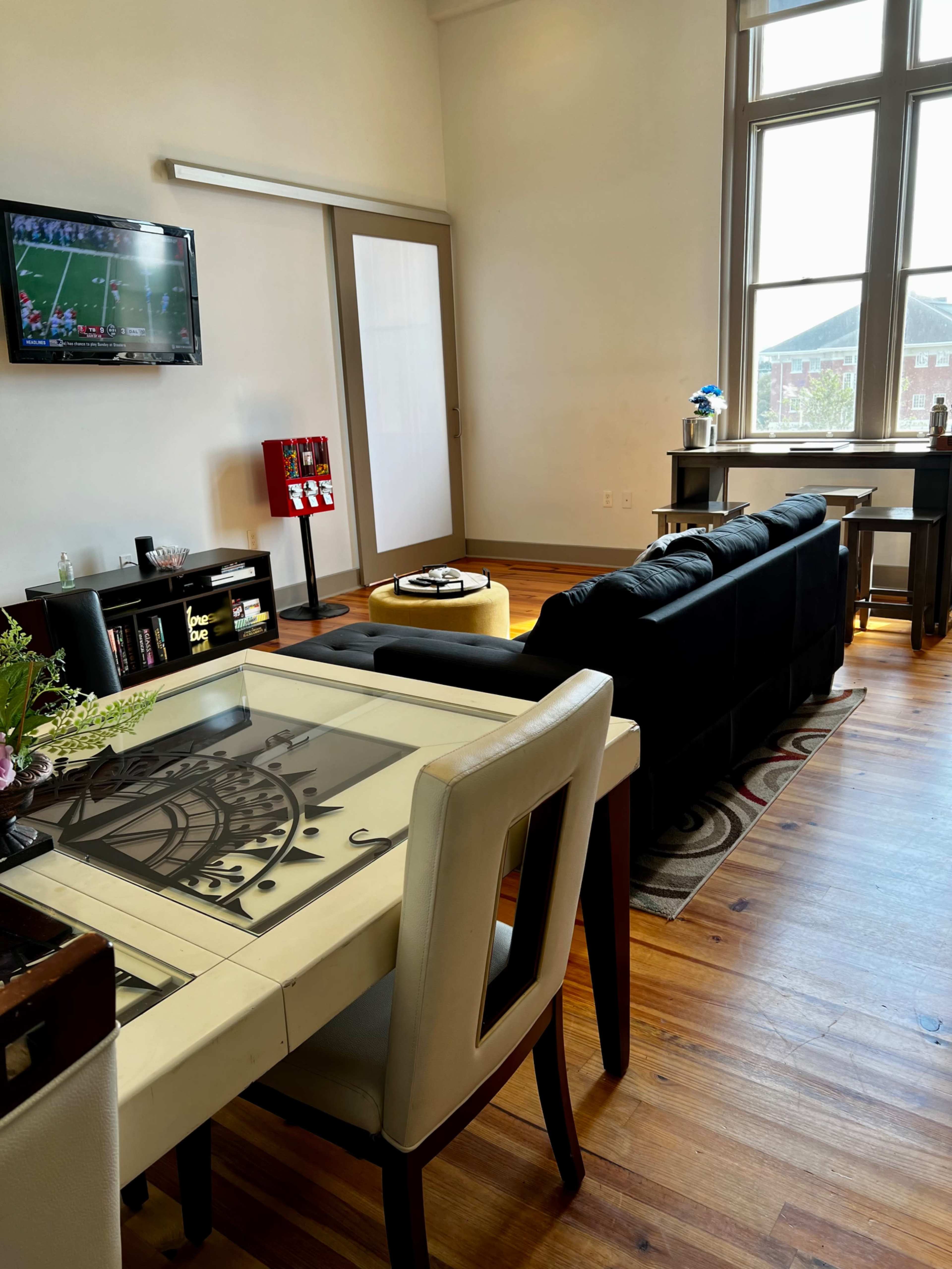 A modern living space features a dining table with a decorative design, a black sofa, a TV displaying a sports game, and a large window illuminating the room.