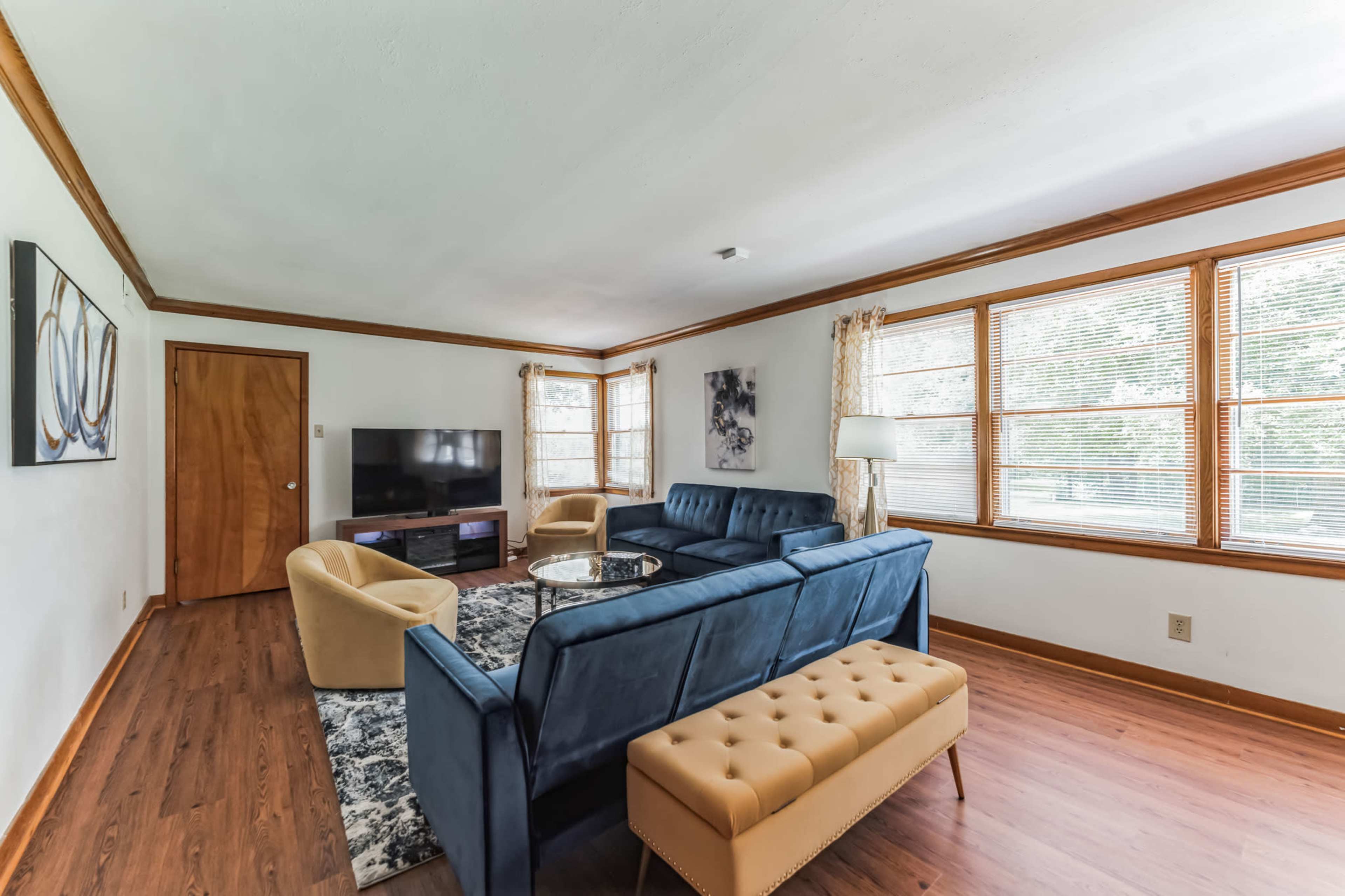 A living room featuring a blue sectional sofa, a beige tufted bench, a flat-screen TV on a stand, and large windows with natural light.