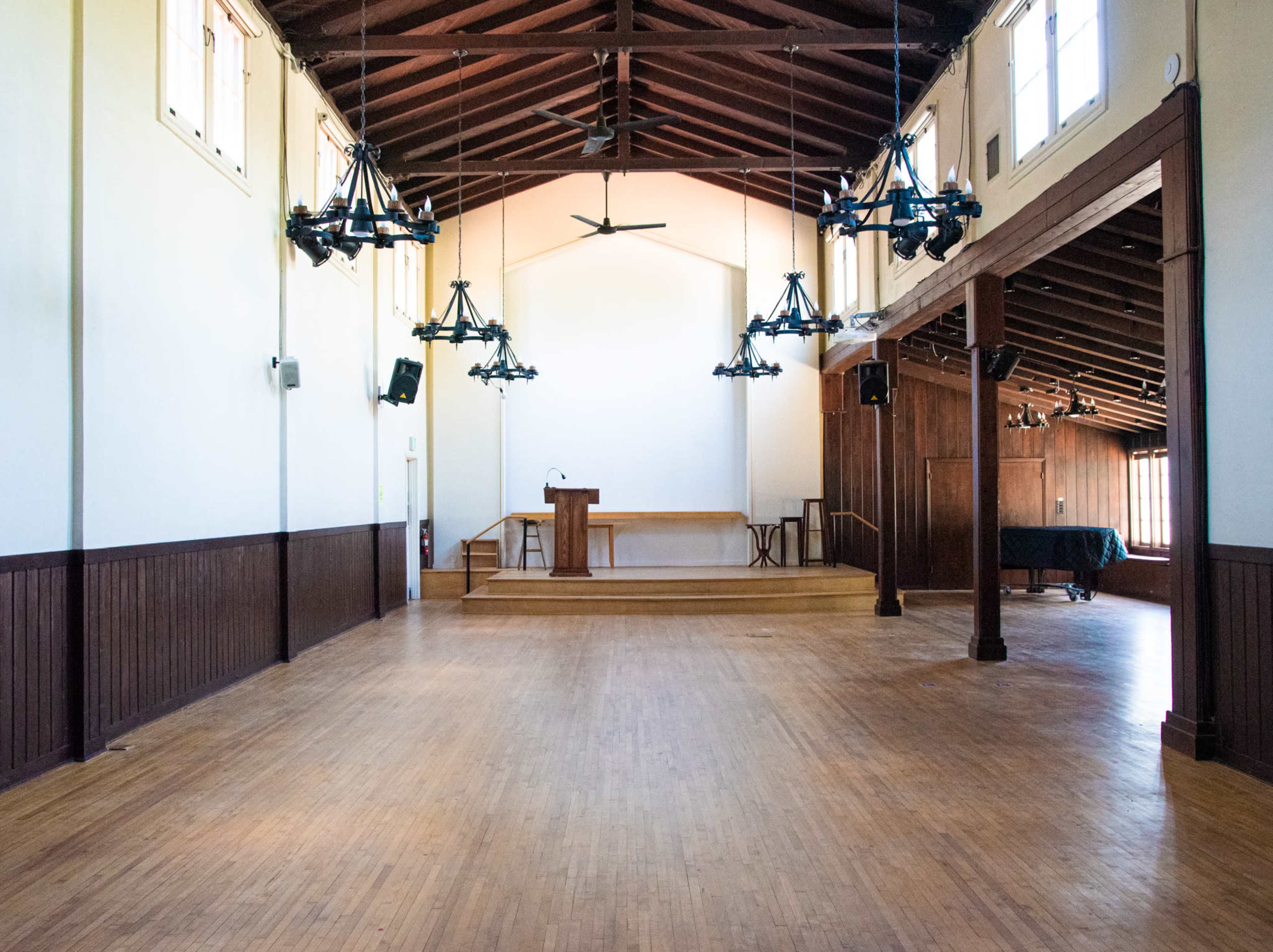 An empty hall with hardwood flooring, large windows, and a stage at one end.