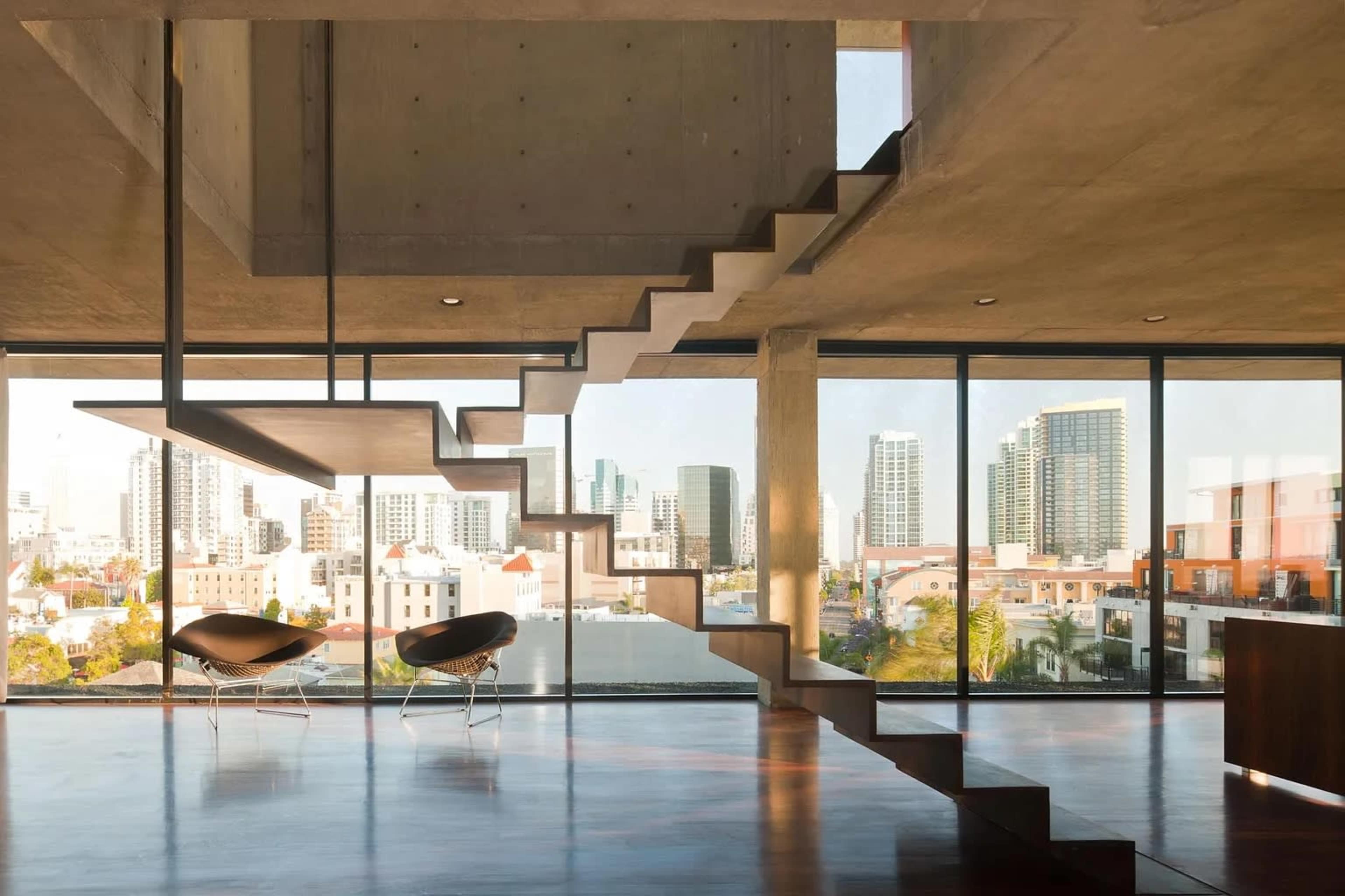 The image shows a modern interior space featuring a minimalist staircase leading to an upper level, with large windows showcasing a city skyline in the background.