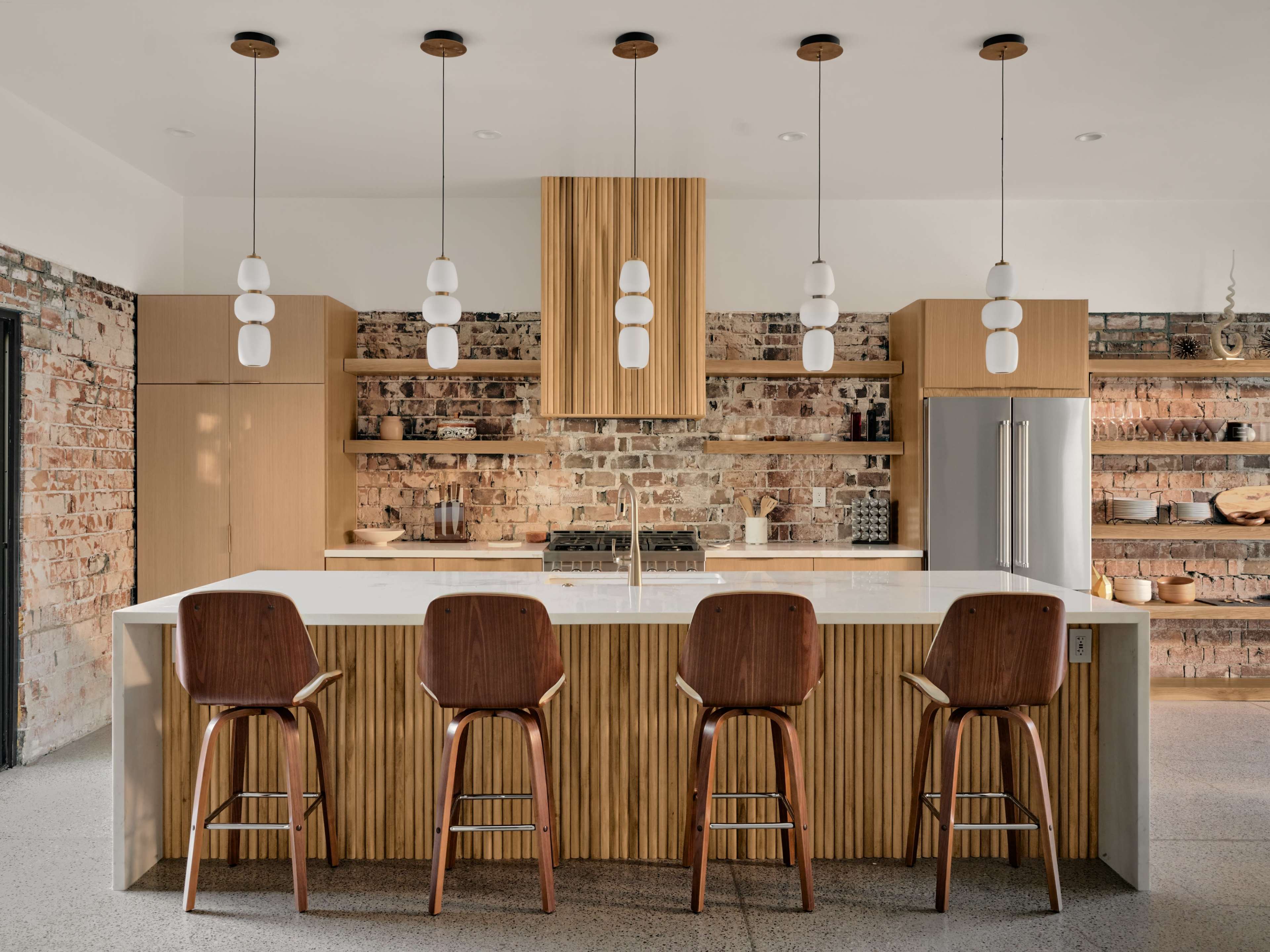 A modern kitchen features a large island with wooden bar stools, pendant lighting, and exposed brick walls.