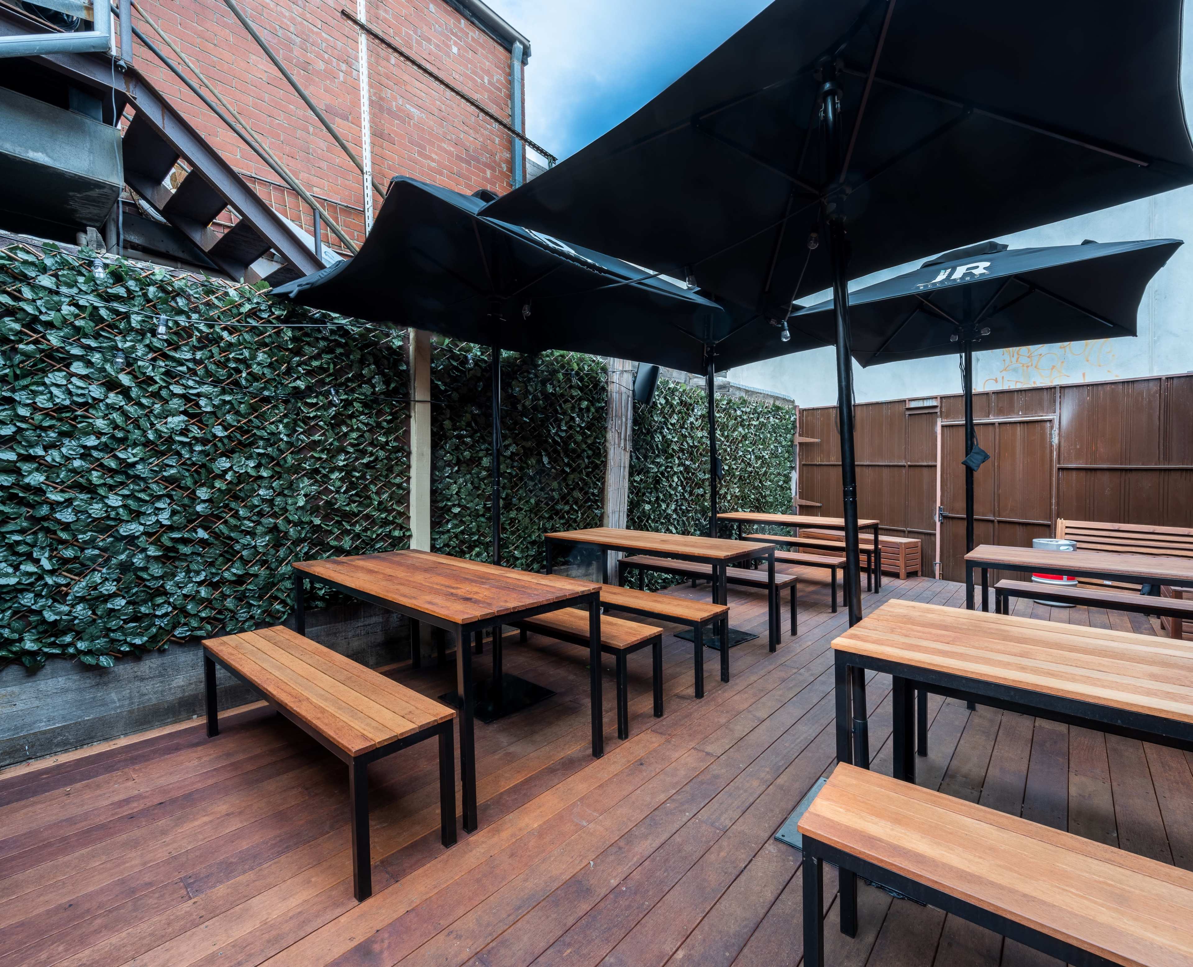 The image shows an outdoor seating area with wooden benches and tables under large black umbrellas, surrounded by a green leafy wall.
