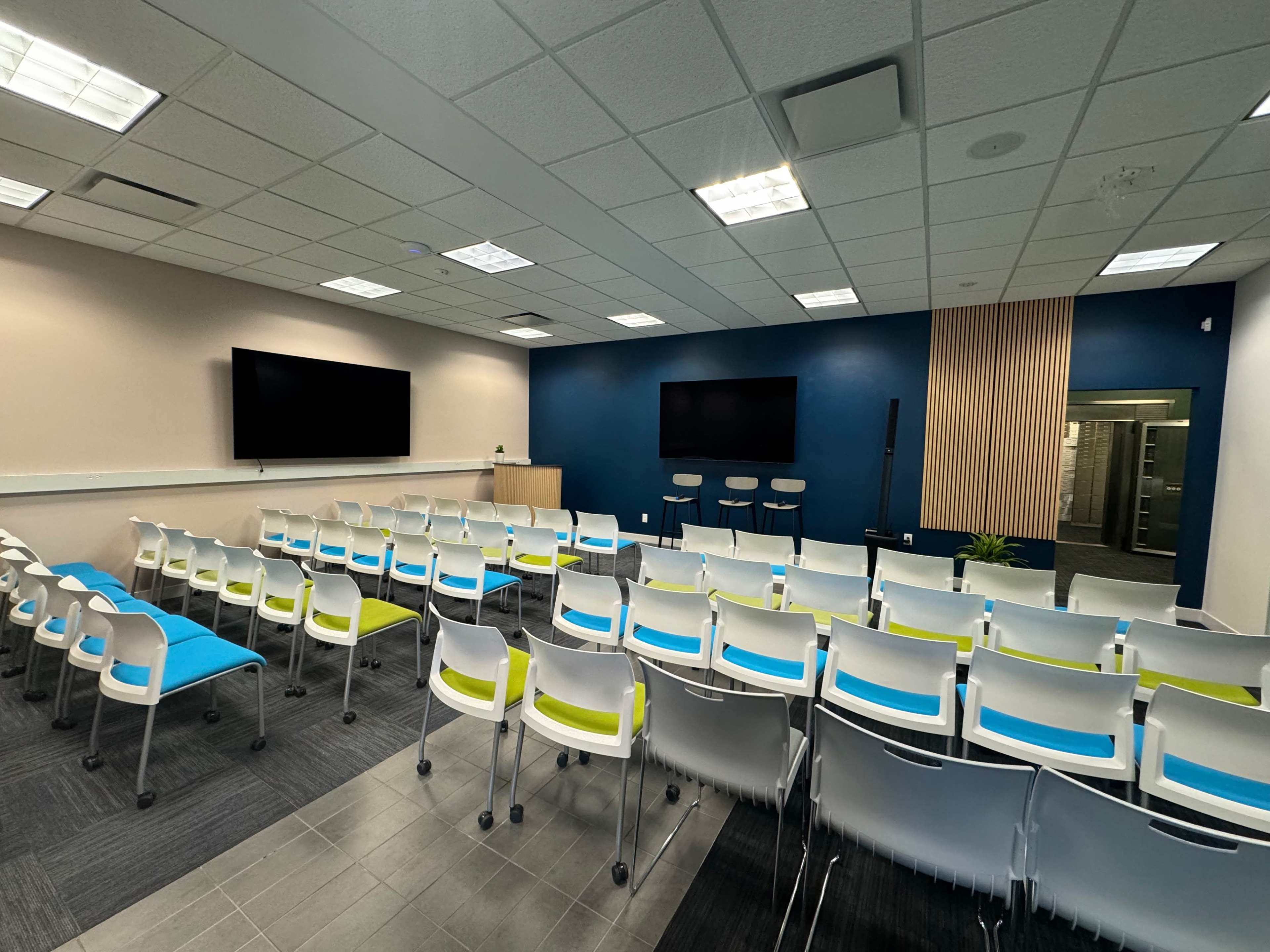 A modern conference room with rows of white chairs featuring blue and green accents, two large screens on the wall, and a backdrop of blue paint and wooden paneling.