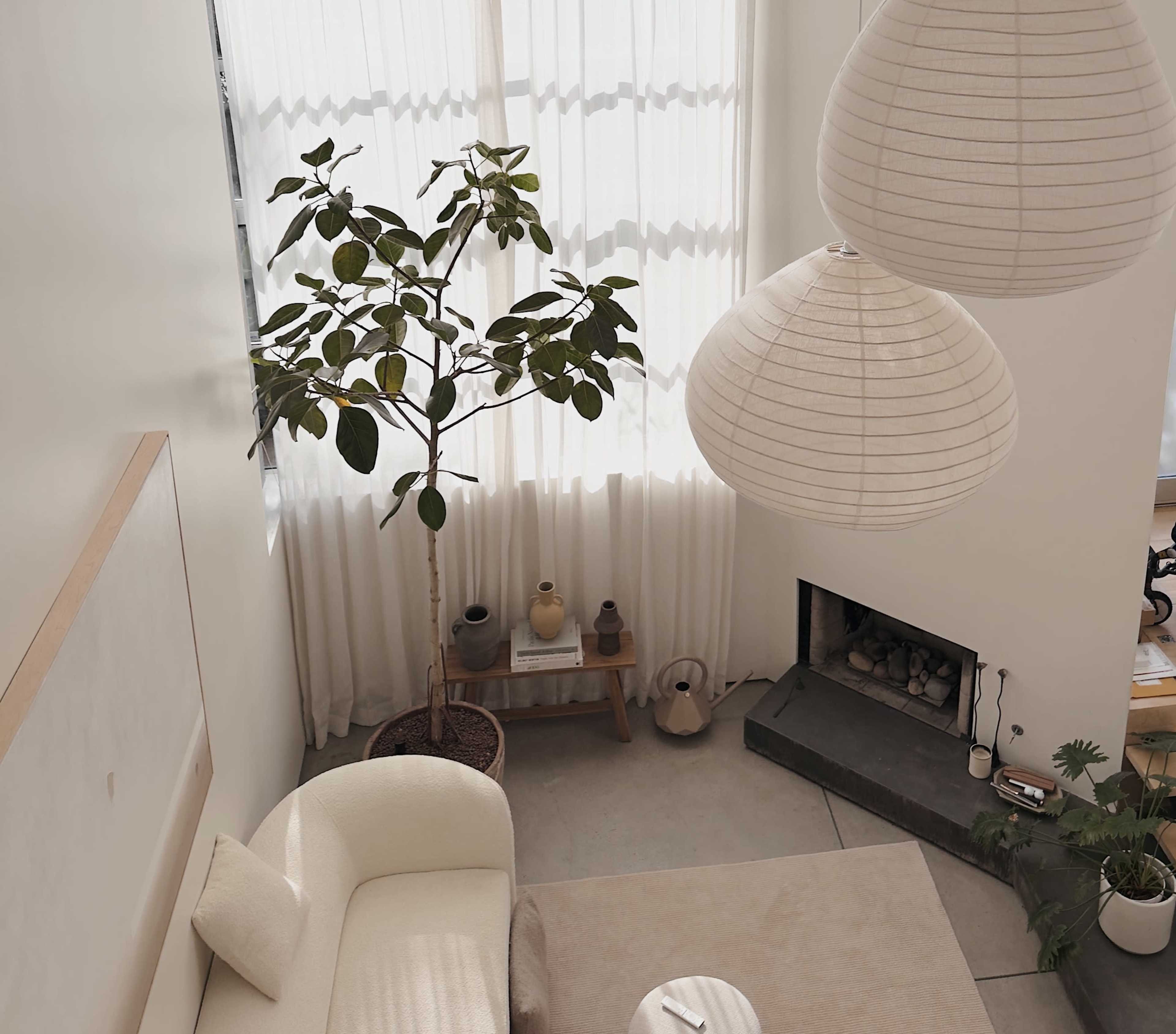 The image shows a modern living room with large windows, a potted plant, and two pendant lamps hanging above a light-colored sofa.