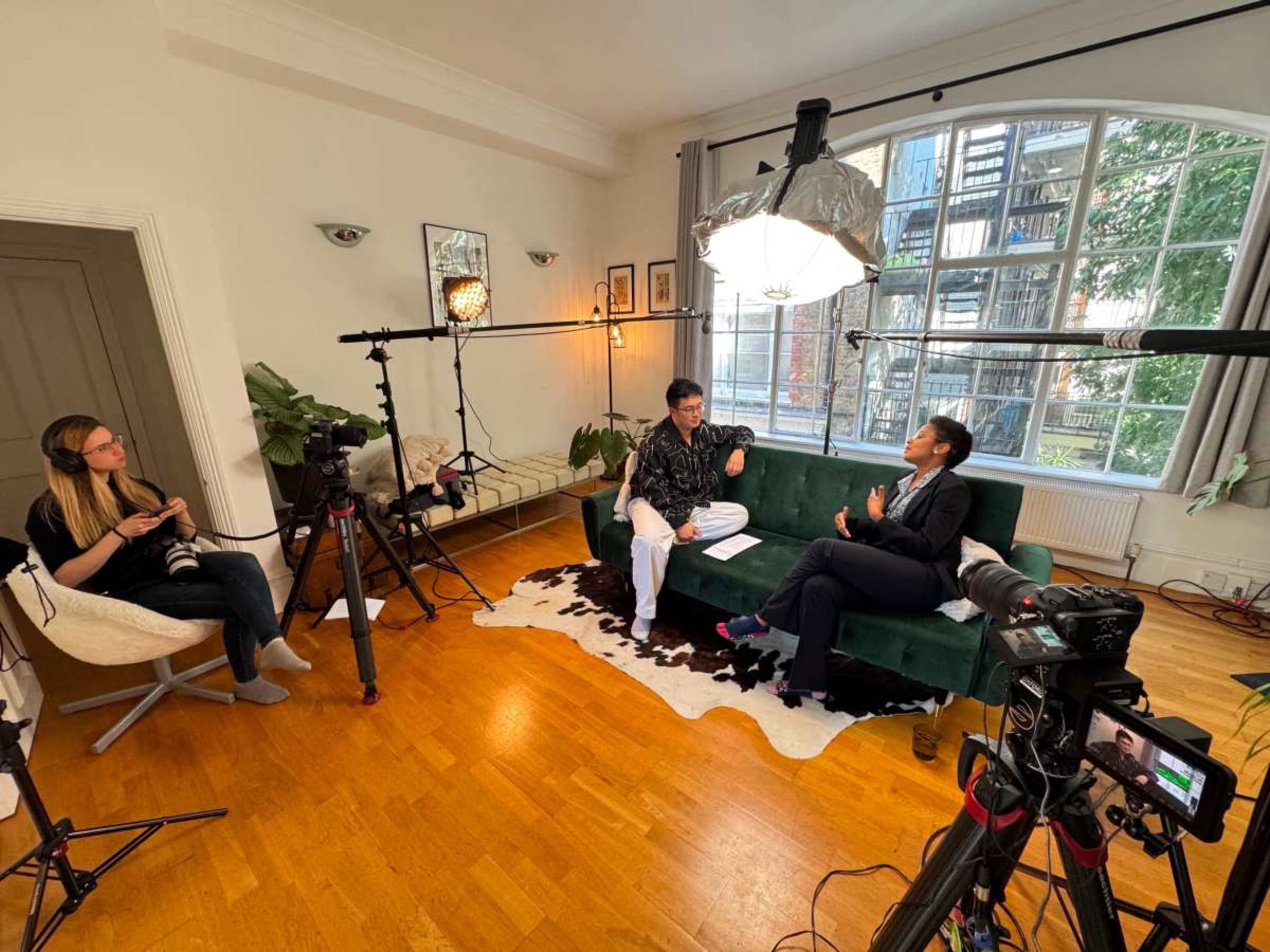 The image shows a two-person interview setup in a well-lit room with a green couch, cameras, and lighting equipment arranged around them.