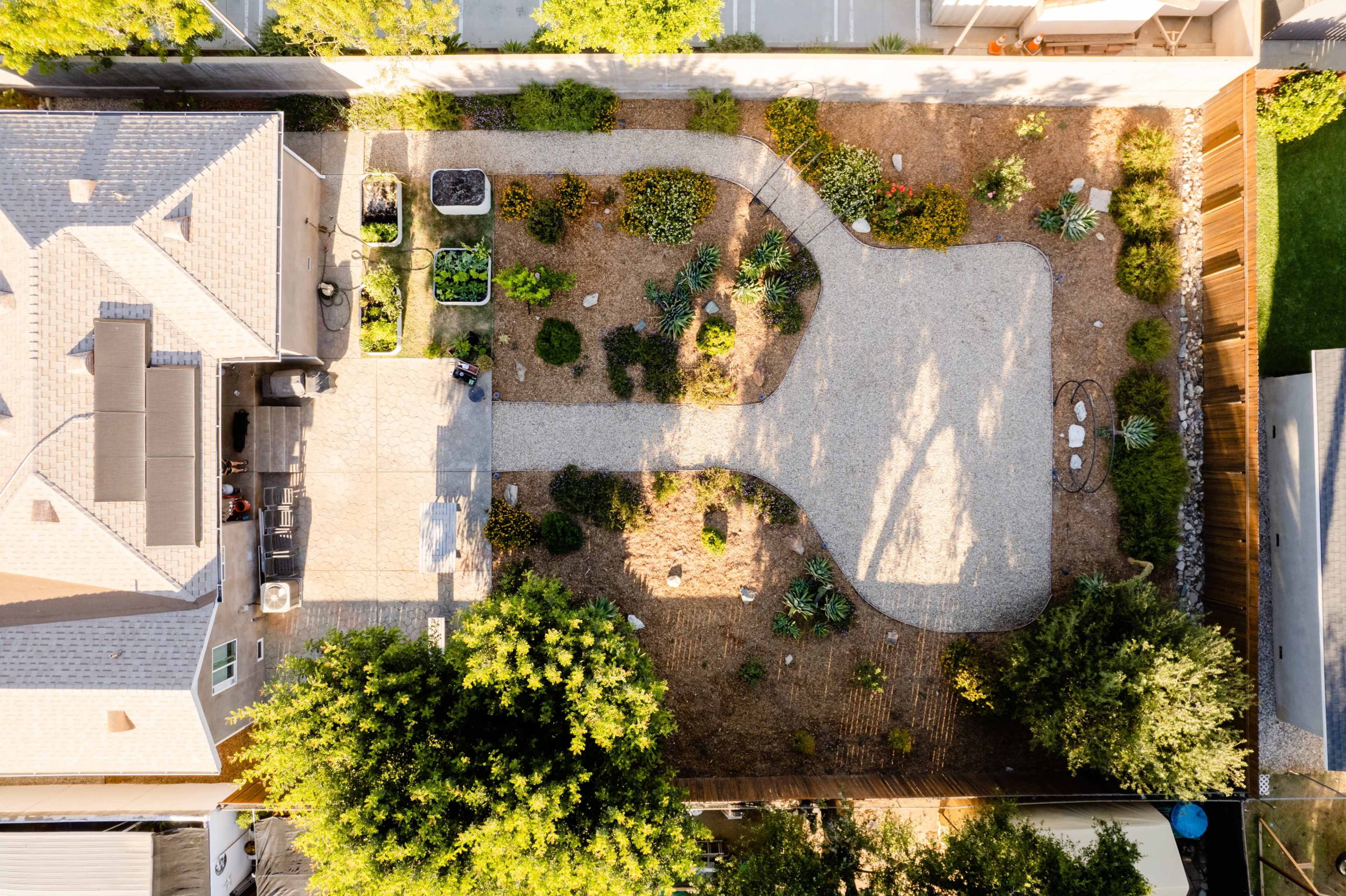 An aerial view shows a landscaped backyard with pathways, garden beds, and various plants surrounding a patio area.