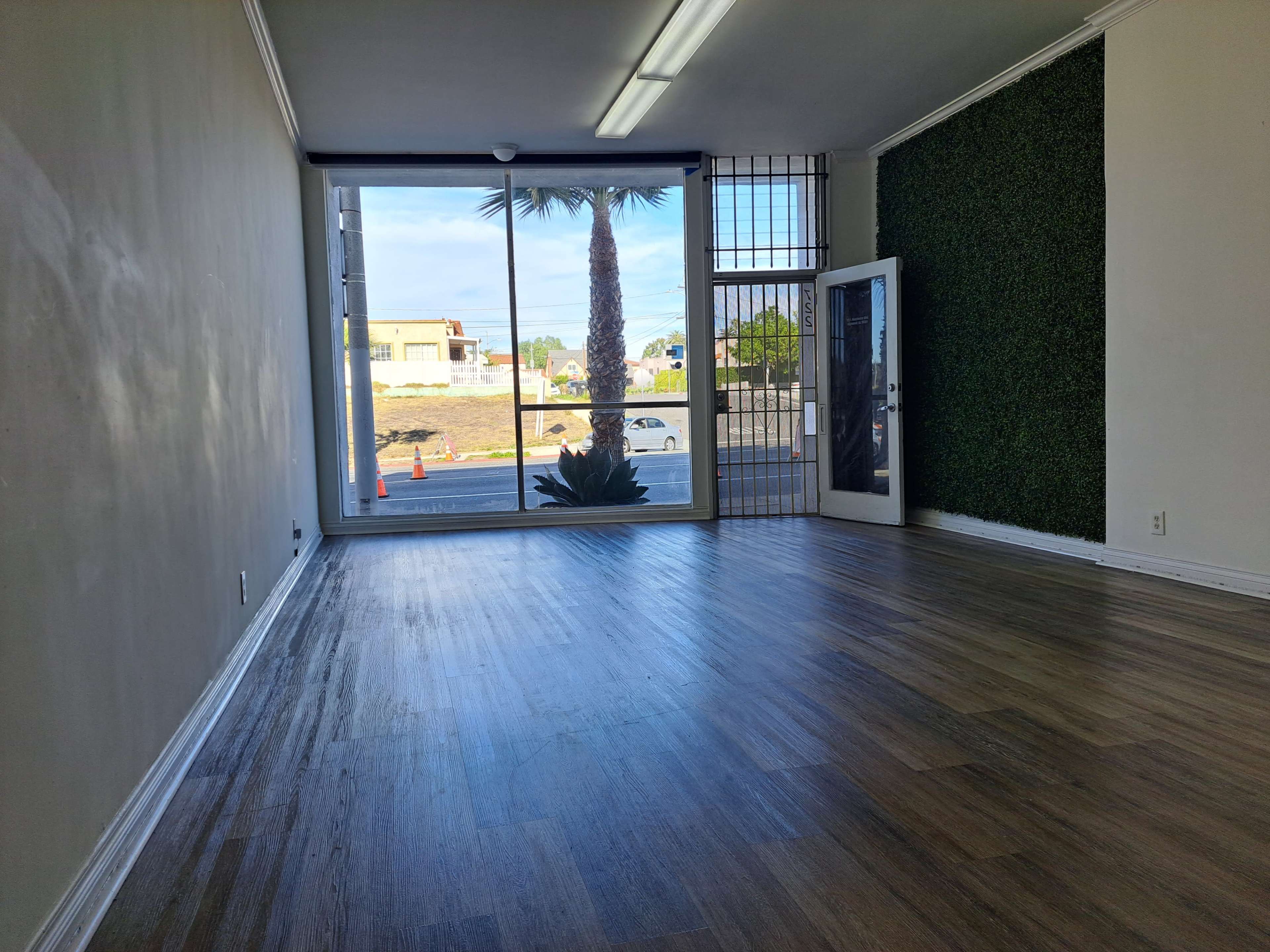 A spacious interior with wooden flooring, featuring a large window that provides a view of a palm tree and an outdoor area.