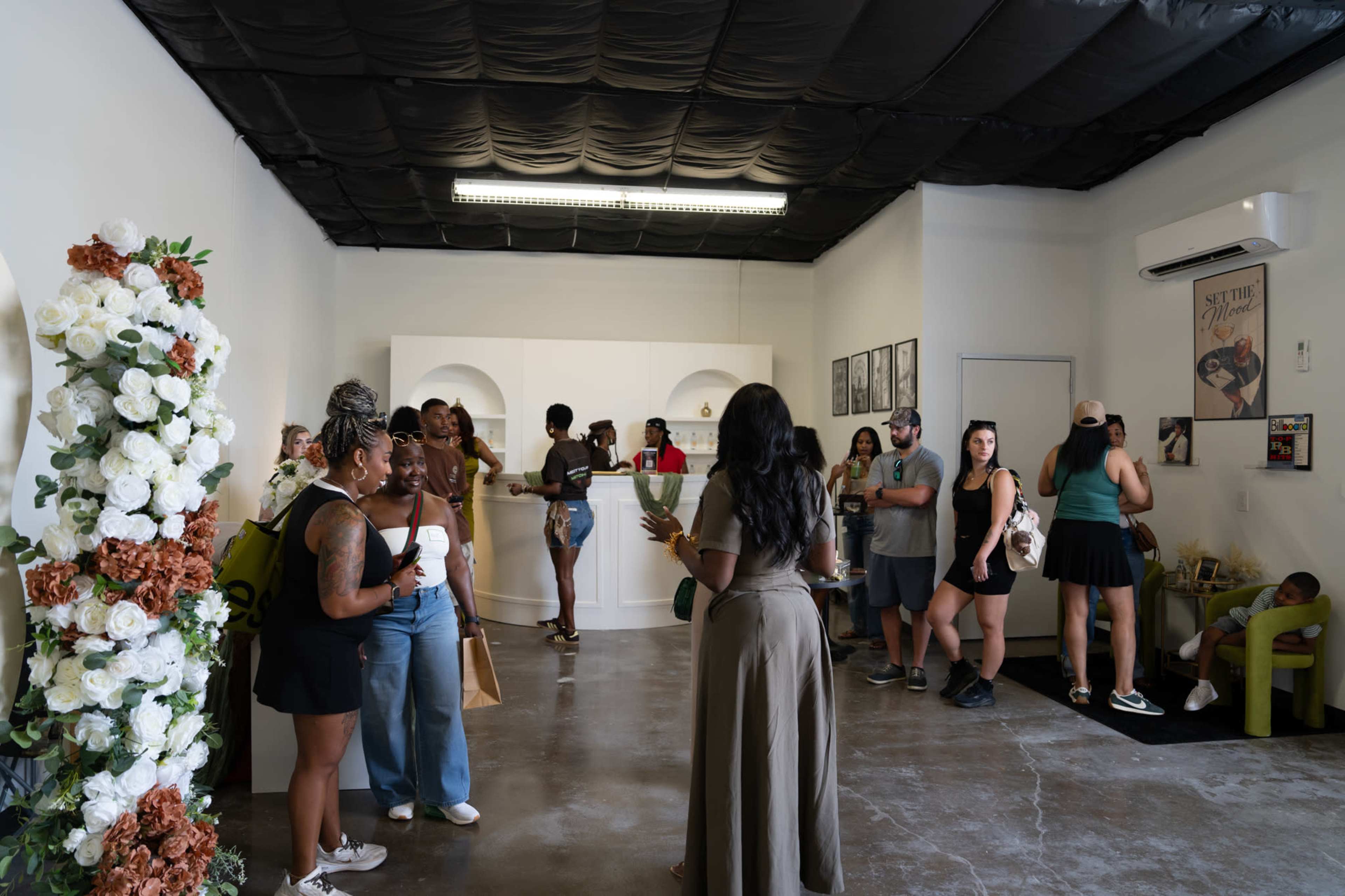 The image shows a group of people socializing in a bright, modern retail space with floral arrangements and shelves displaying products.