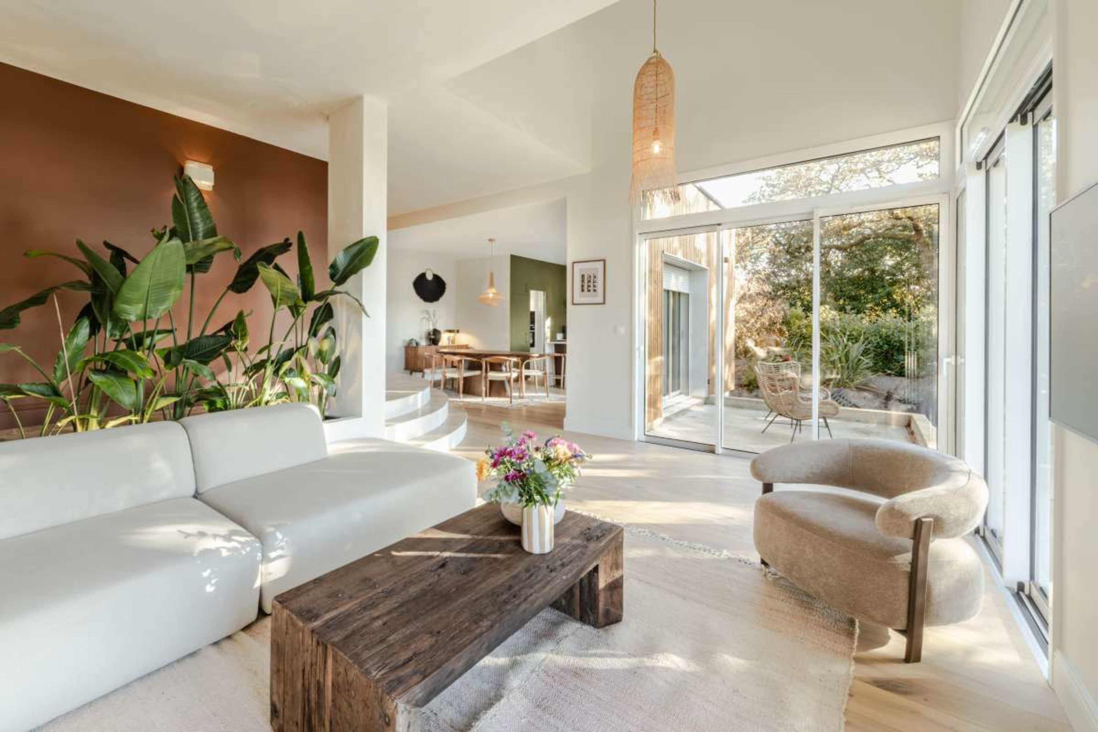 The scene depicts a modern living area featuring a light-colored sofa, a wooden coffee table, and large glass doors opening to a garden.