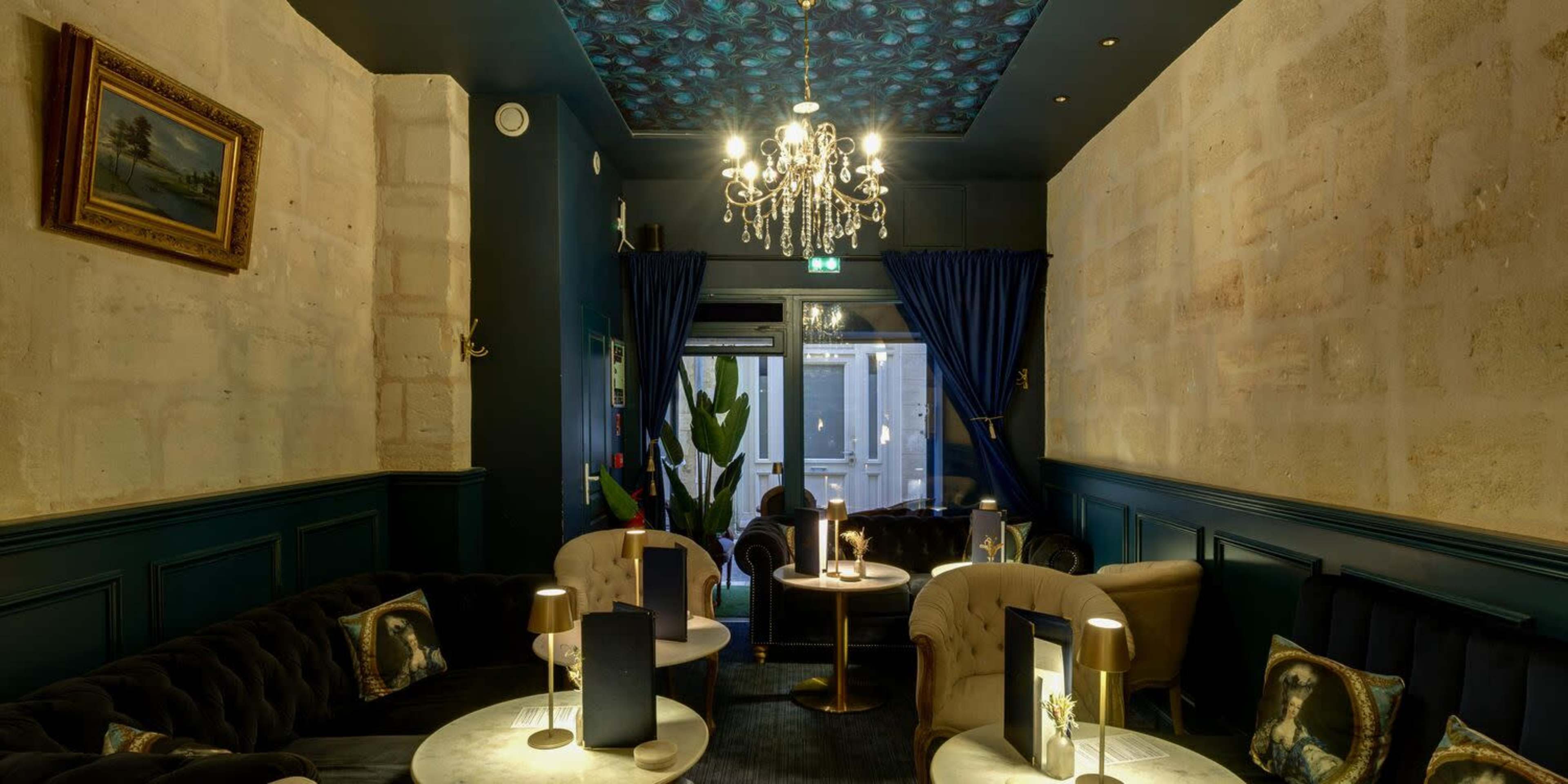 The image shows a cozy lounge area featuring dark upholstered furniture, a chandelier, and artwork on the walls, illuminated by soft lighting.
