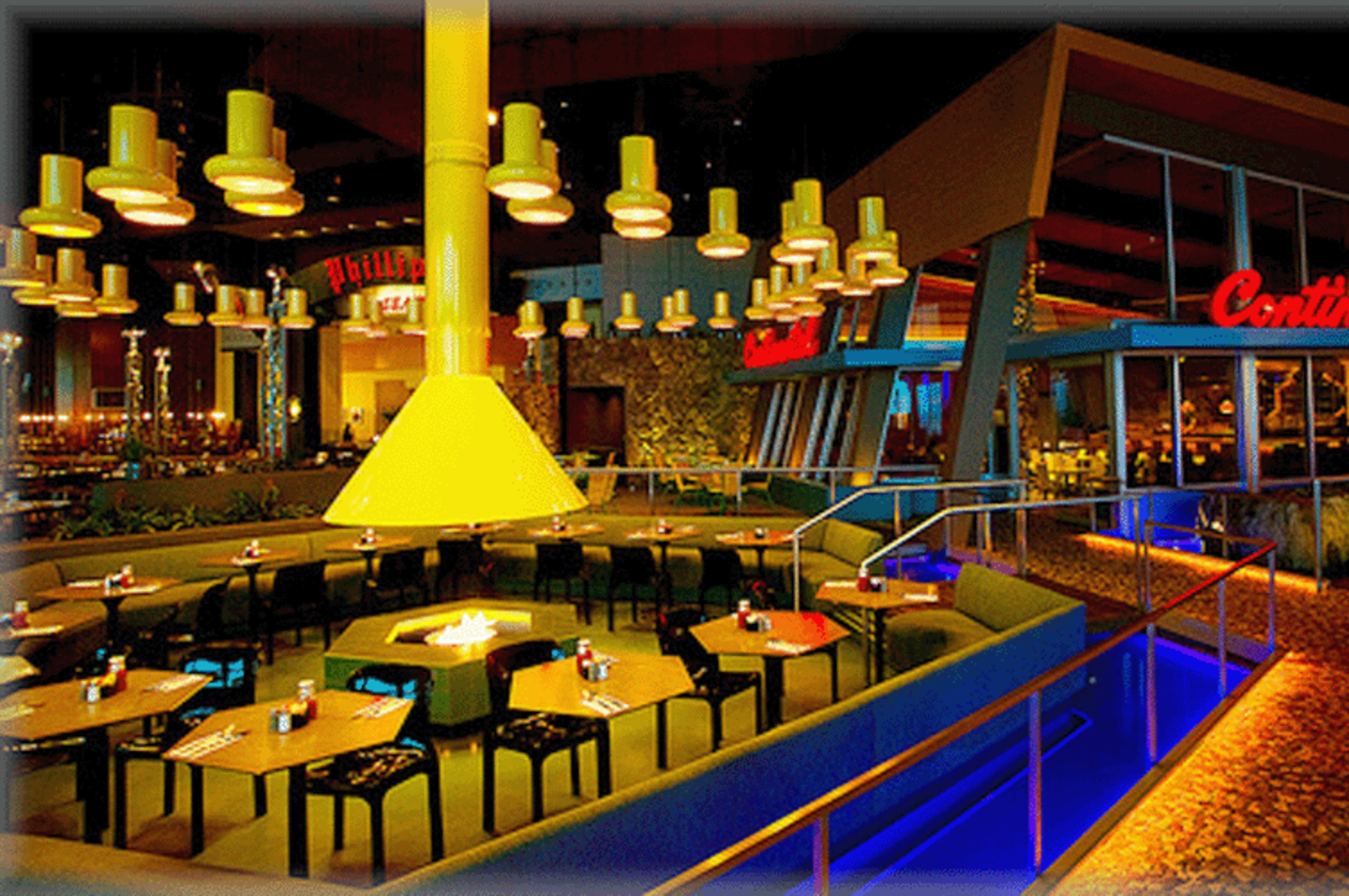 A modern restaurant interior features yellow lighting, a central chimney-like structure, and tables arranged around a fire pit.