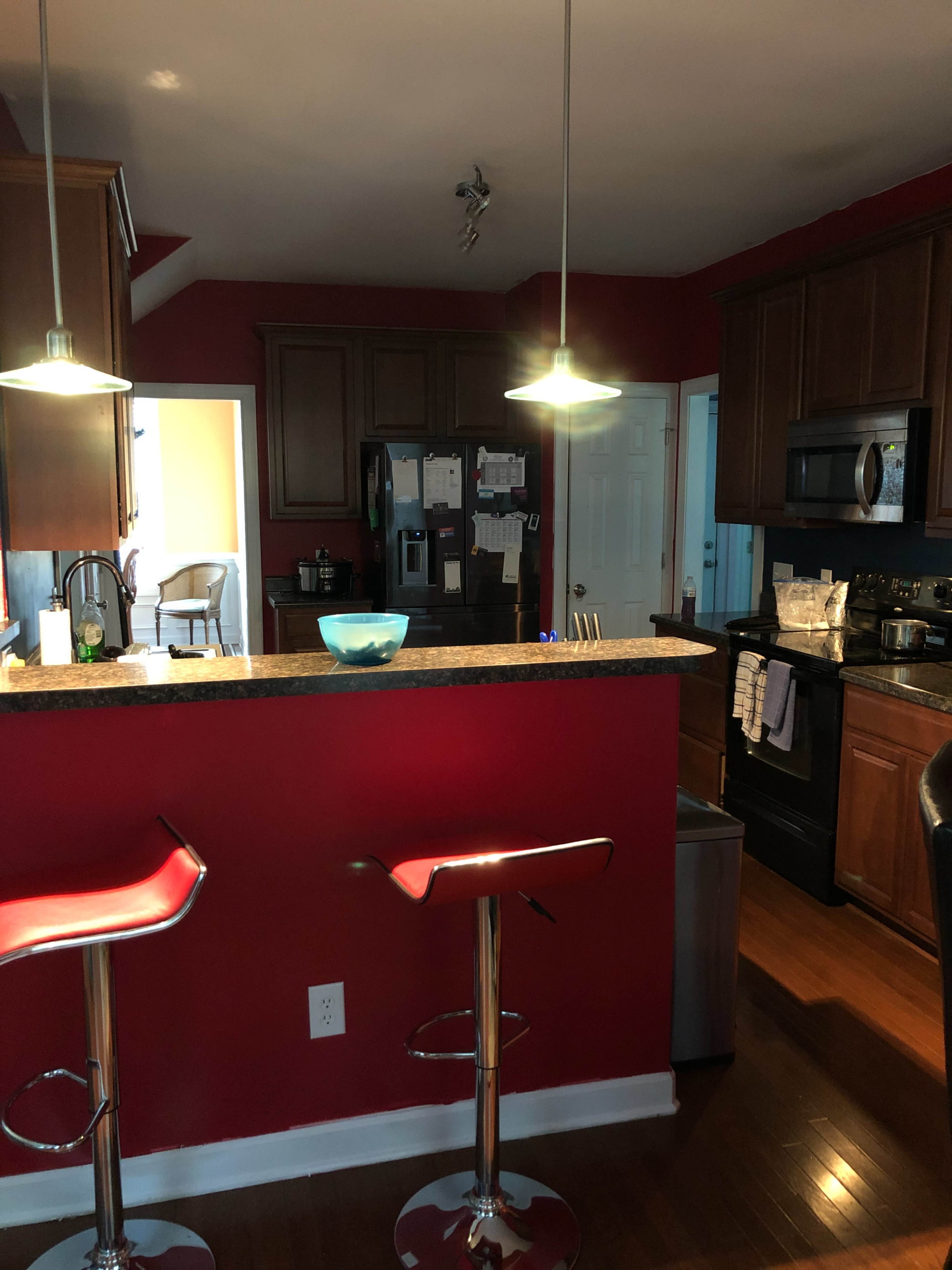 A modern kitchen features a bar with two red stools, dark wooden cabinets, and stainless steel appliances, illuminated by pendant lights.