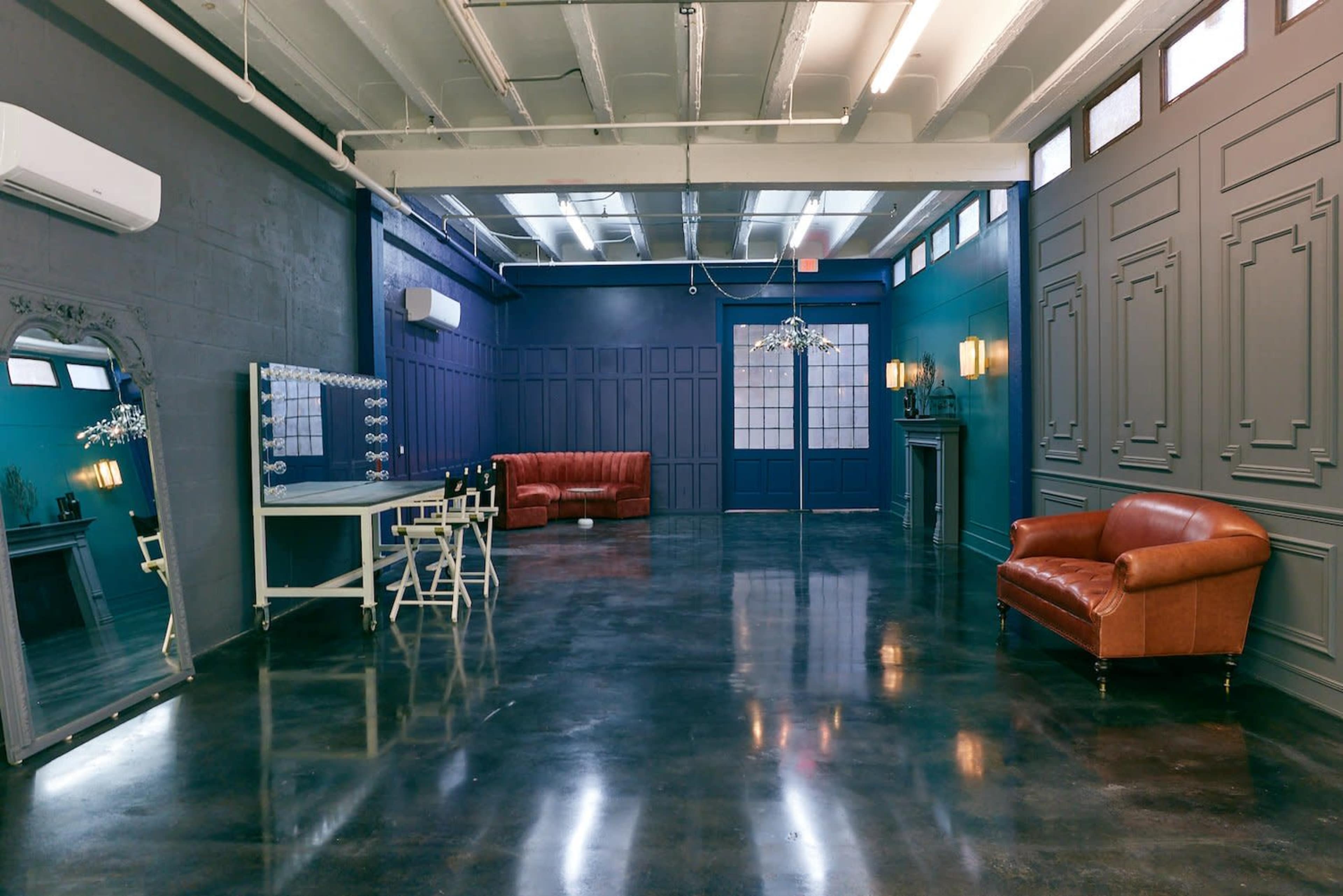 The image shows a spacious room with polished concrete floors, blue and gray walls, a large mirror, a vanity table, two leather sofas, and stylish lighting fixtures.