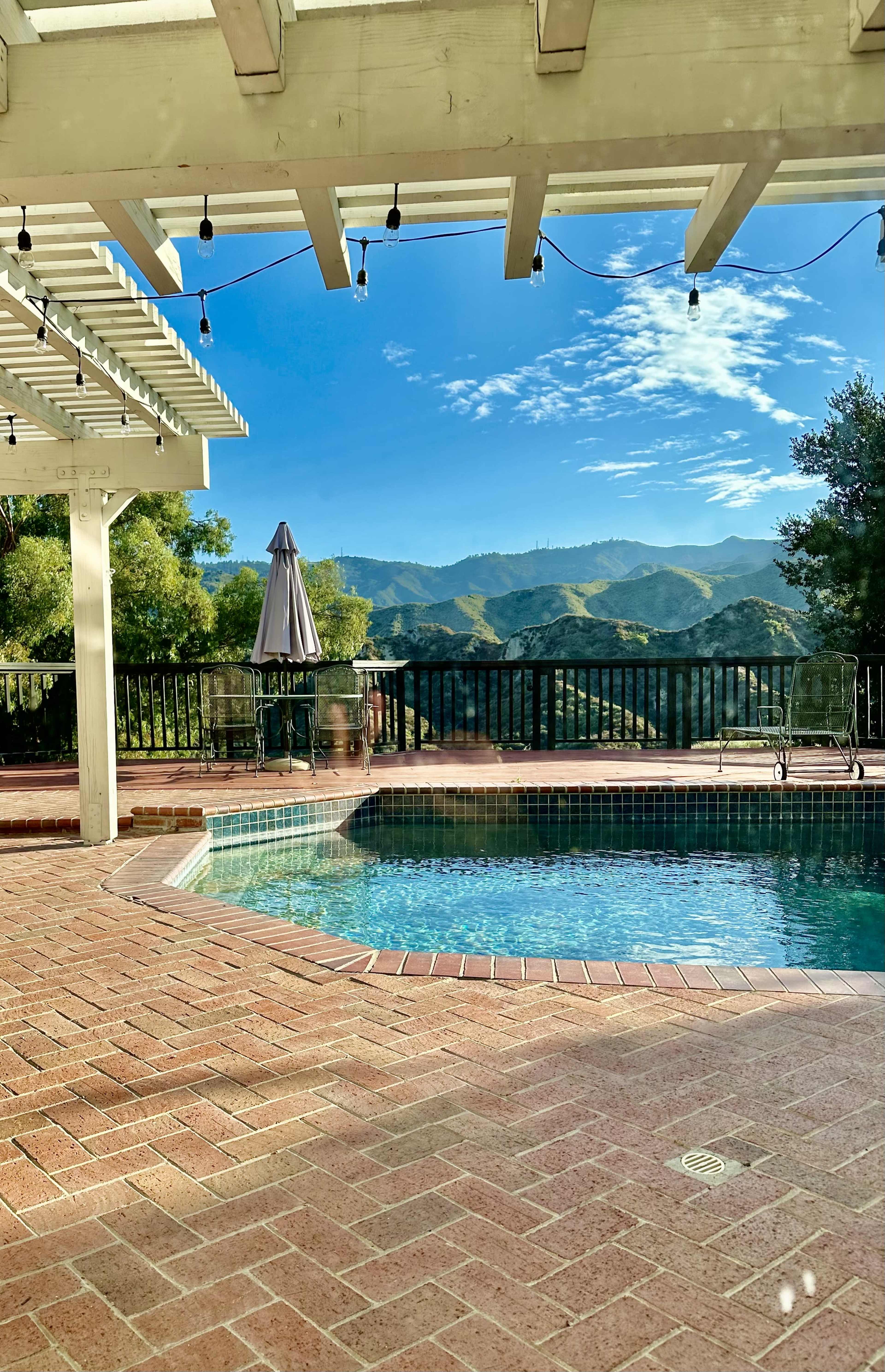 A swimming pool is situated in a patio surrounded by mountains and a blue sky, with a pergola overhead and string lights hanging above.