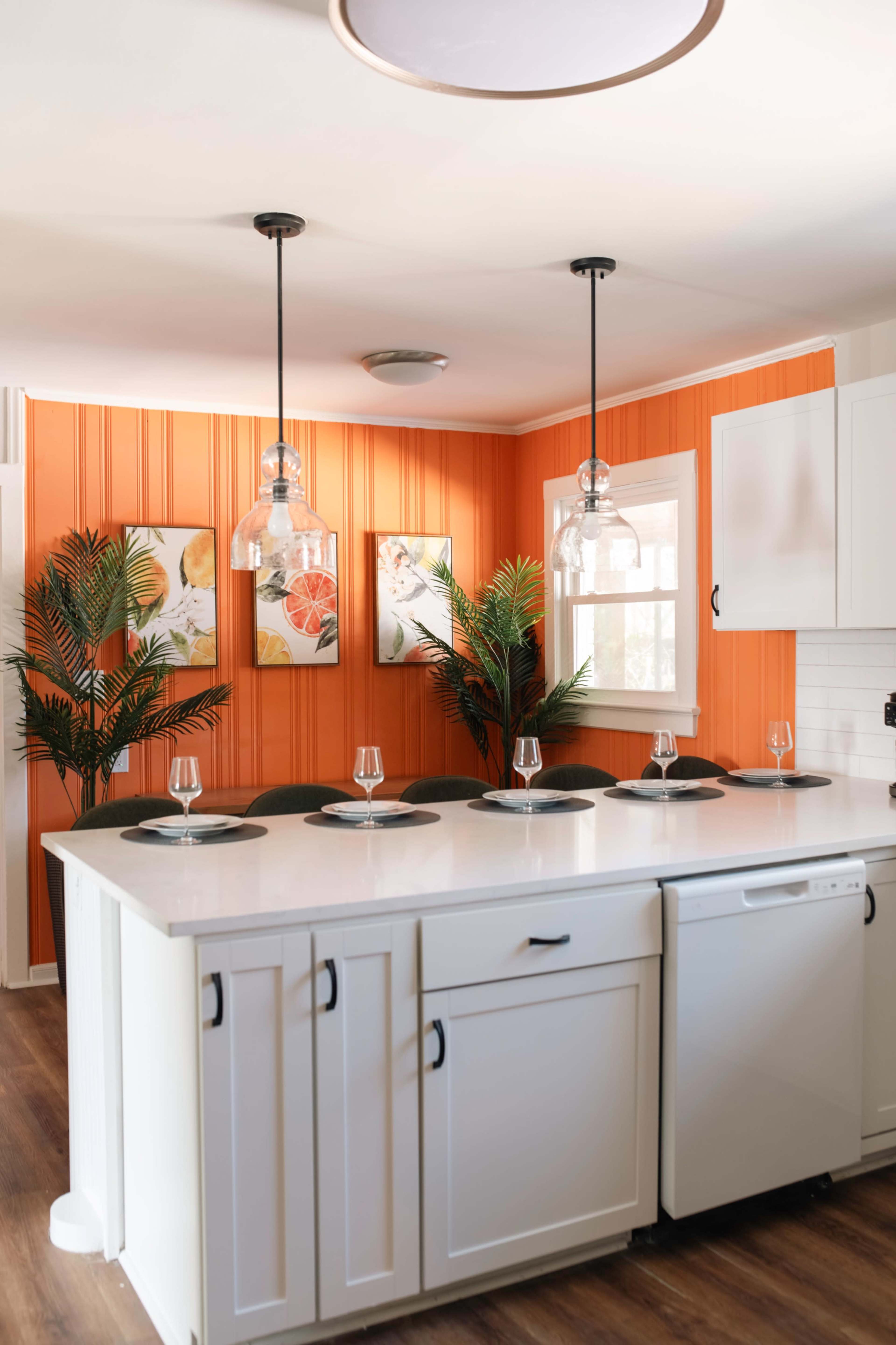 The kitchen features a white island with bar seating, surrounded by vibrant orange walls and framed floral artwork.