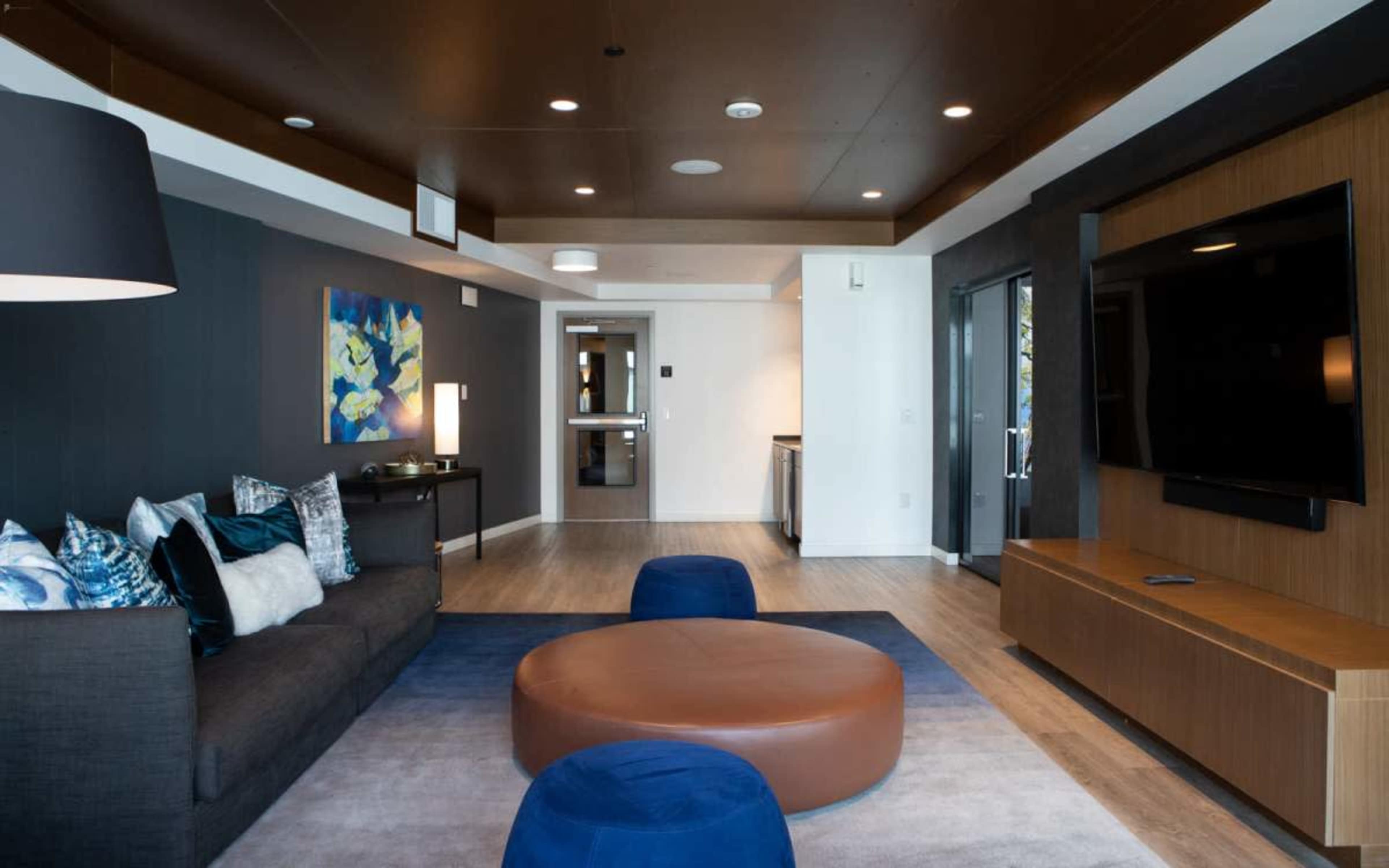 The image shows a modern living room featuring a dark-colored sofa, a circular brown coffee table, blue accent chairs, a mounted television, and a door leading to another room.