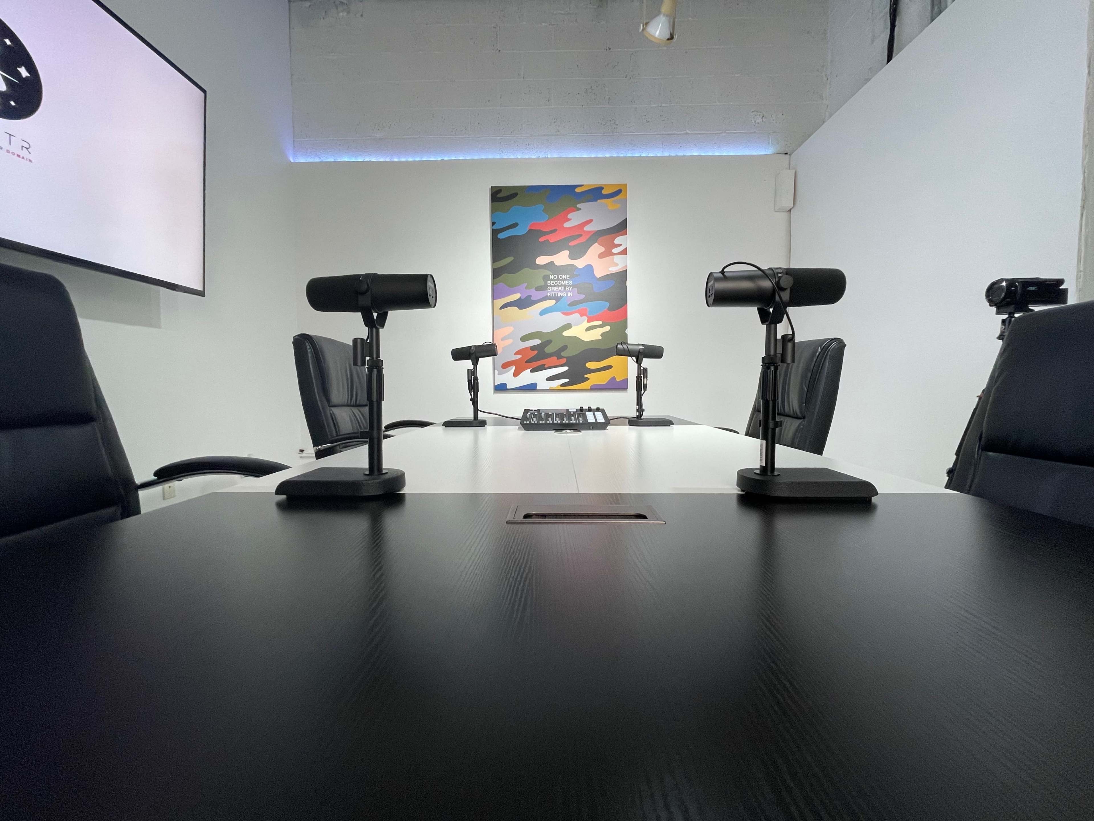 A conference room features a round table with several microphones and black leather chairs, set against a colorful abstract wall mural.