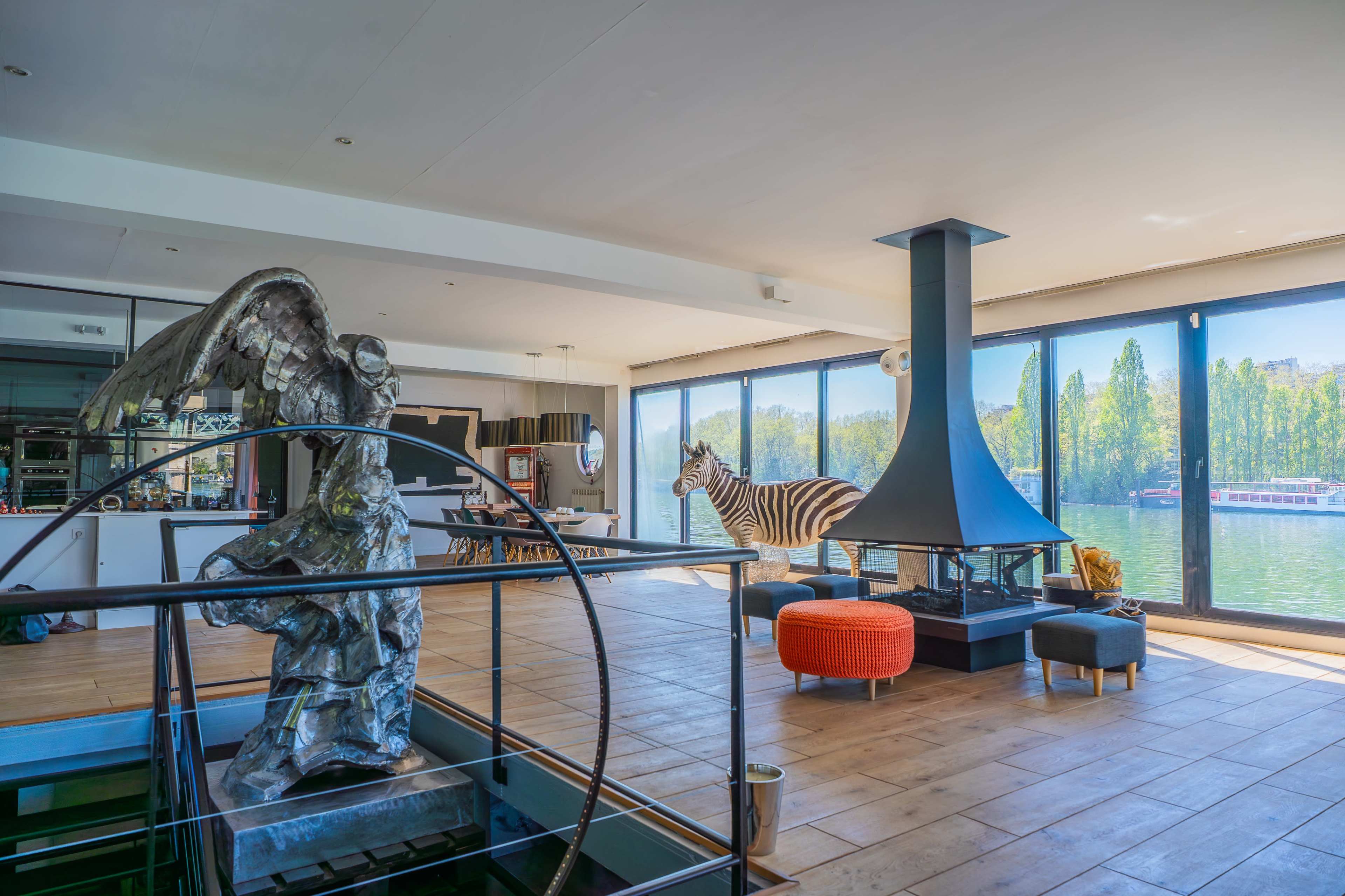 The interior space features large windows overlooking a body of water, a central fireplace, a zebra statue, and an angel sculpture.