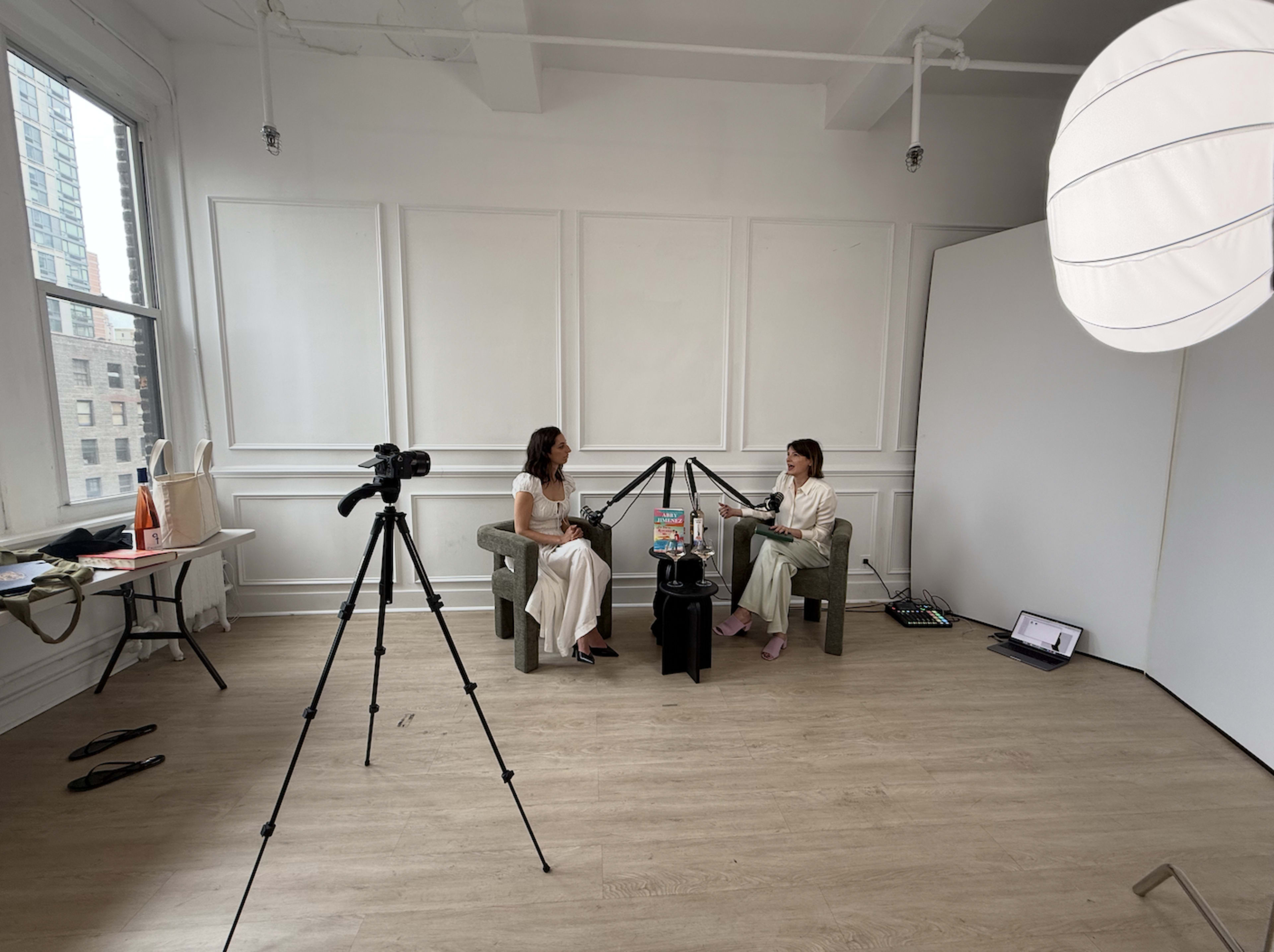 Two women sit in a minimalist studio with a camera and tripod set up for a video recording.