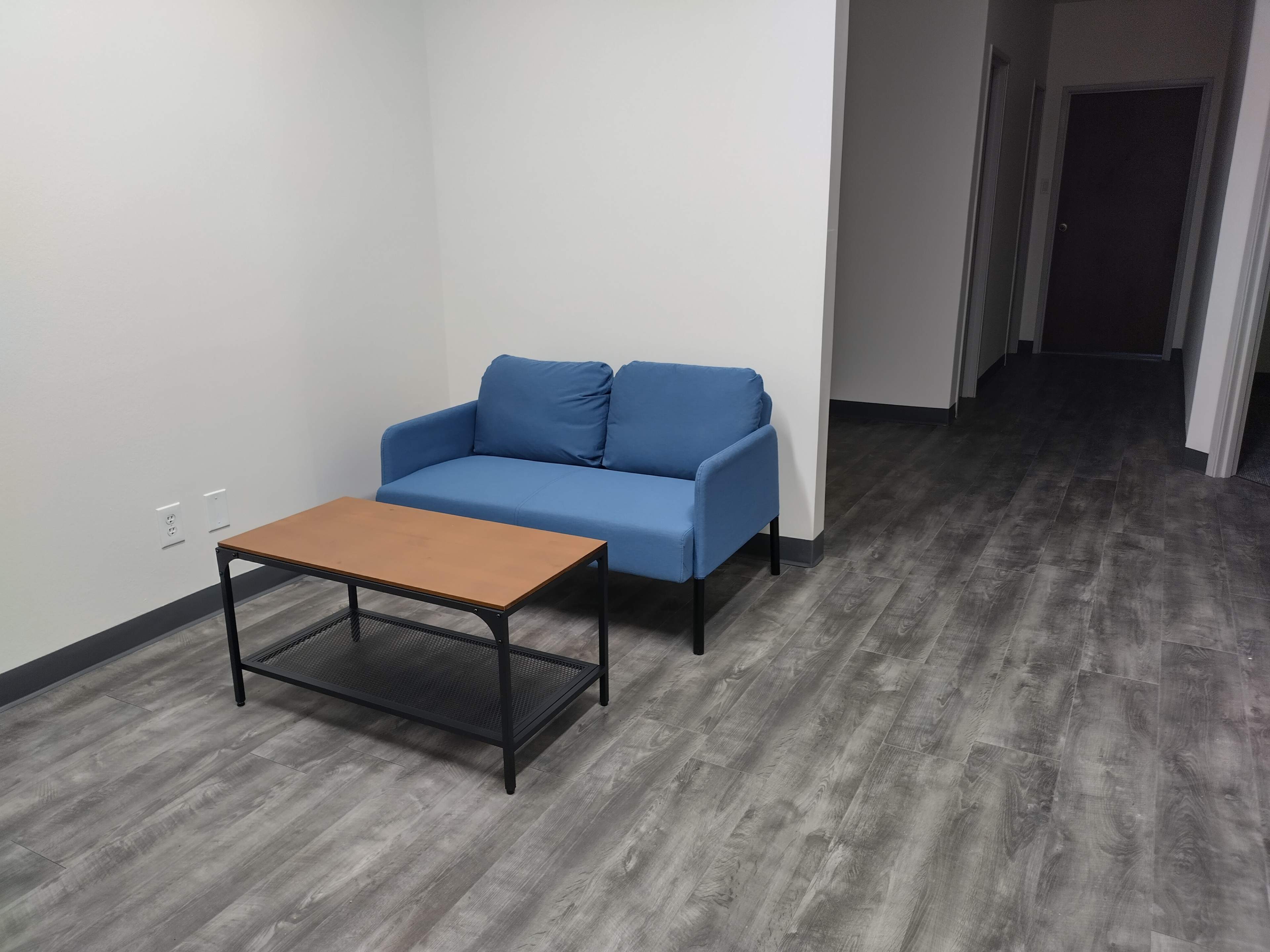 The image shows a blue couch and a wooden coffee table in a sparsely furnished office space with gray flooring and a hallway visible in the background.