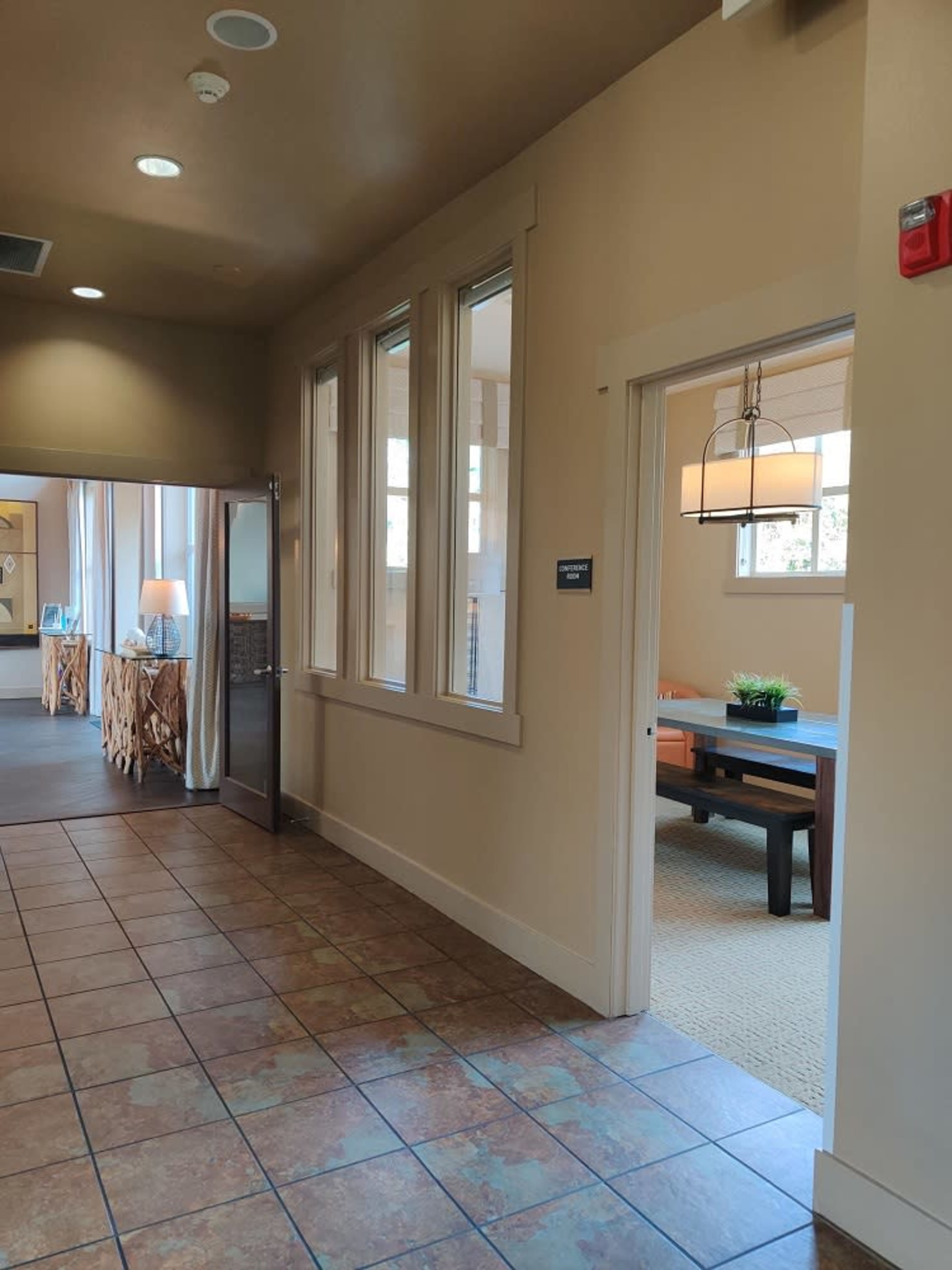 The image shows a hallway with tiled flooring and a doorway leading to a room with a table and plants.