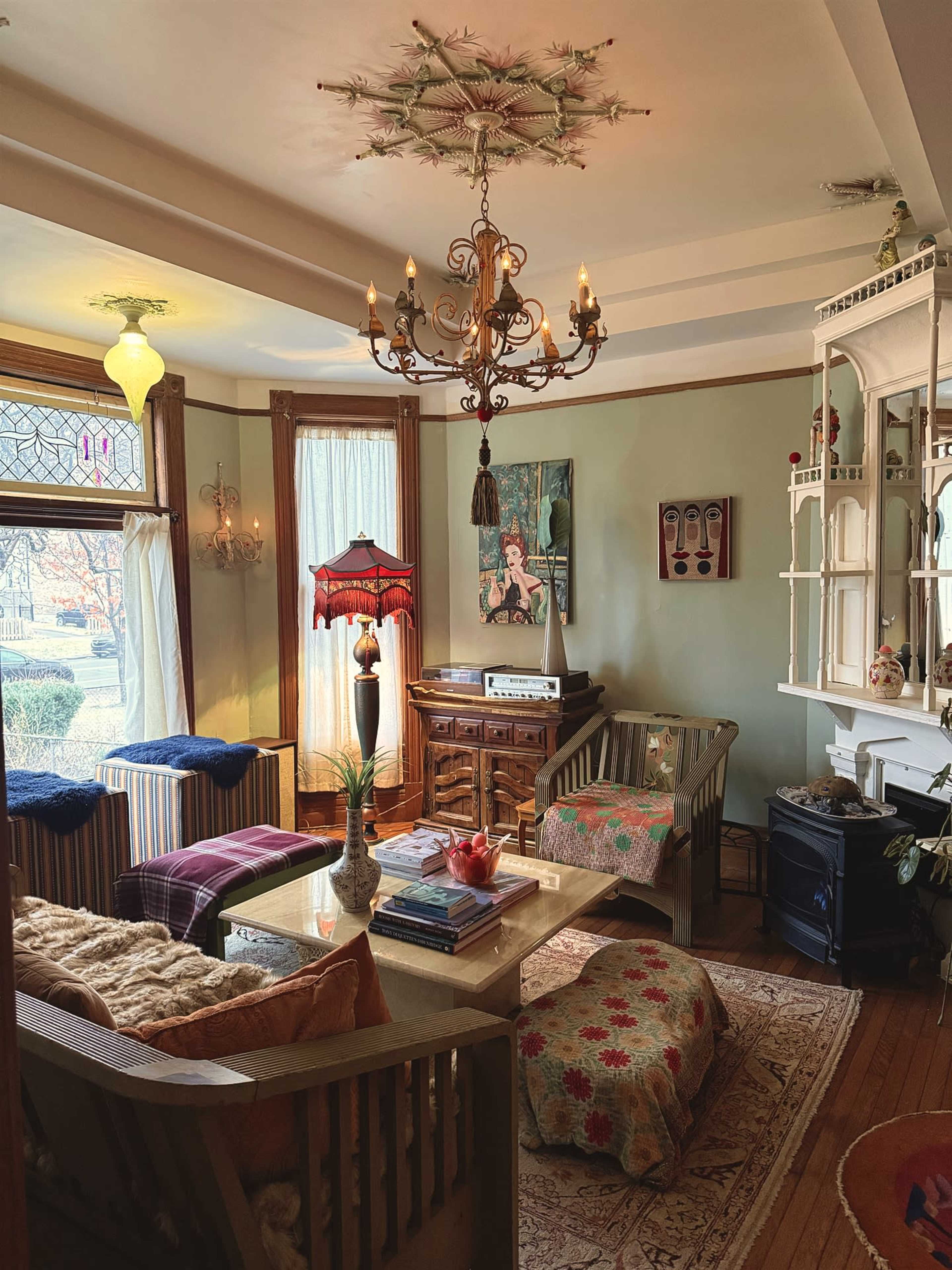 A cozy living room features vintage furniture, a chandelier, and large windows that allow natural light to illuminate the space.