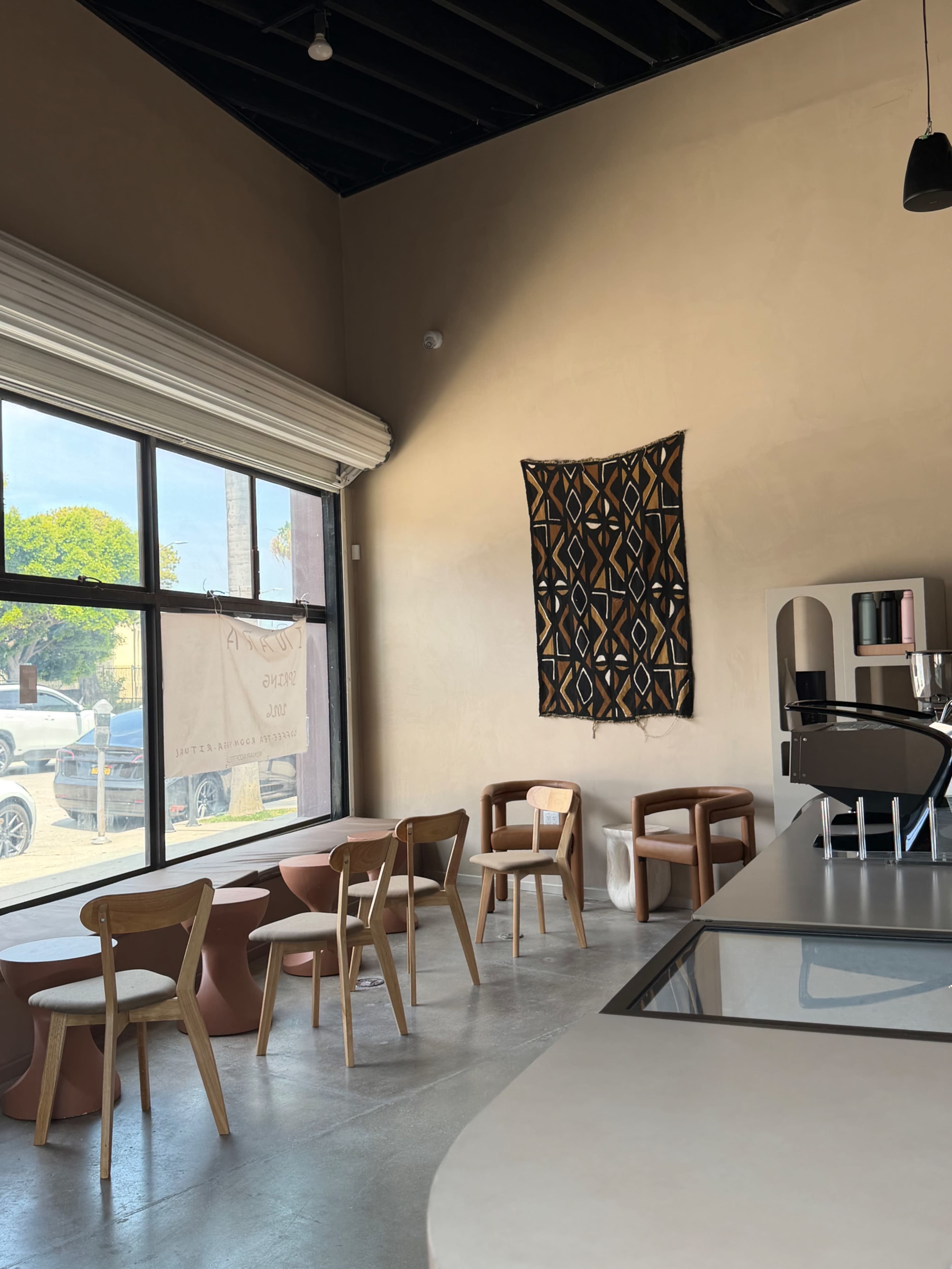 The image shows a minimalist café interior with wooden chairs, a simple counter, and a textile hanging on the wall, along with large windows letting in natural light.