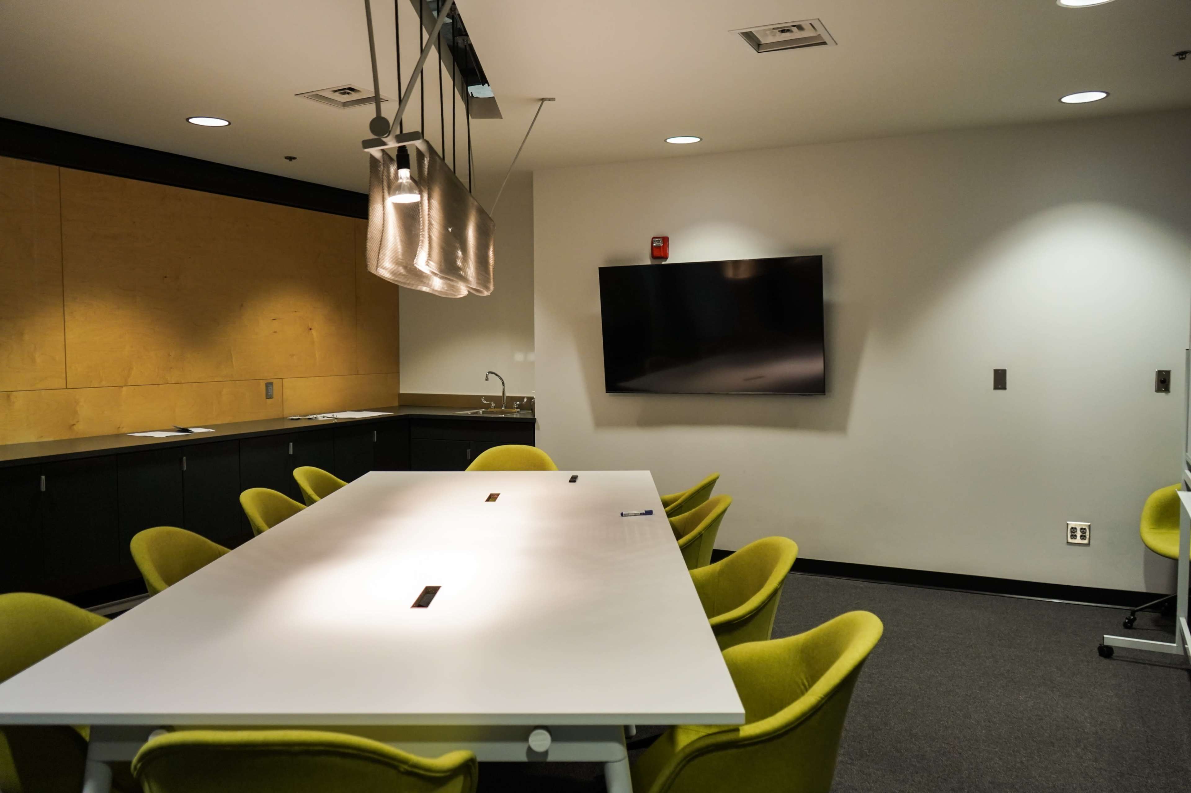 A conference room features a large white table surrounded by green chairs, with a wall-mounted television and a kitchenette in the background.