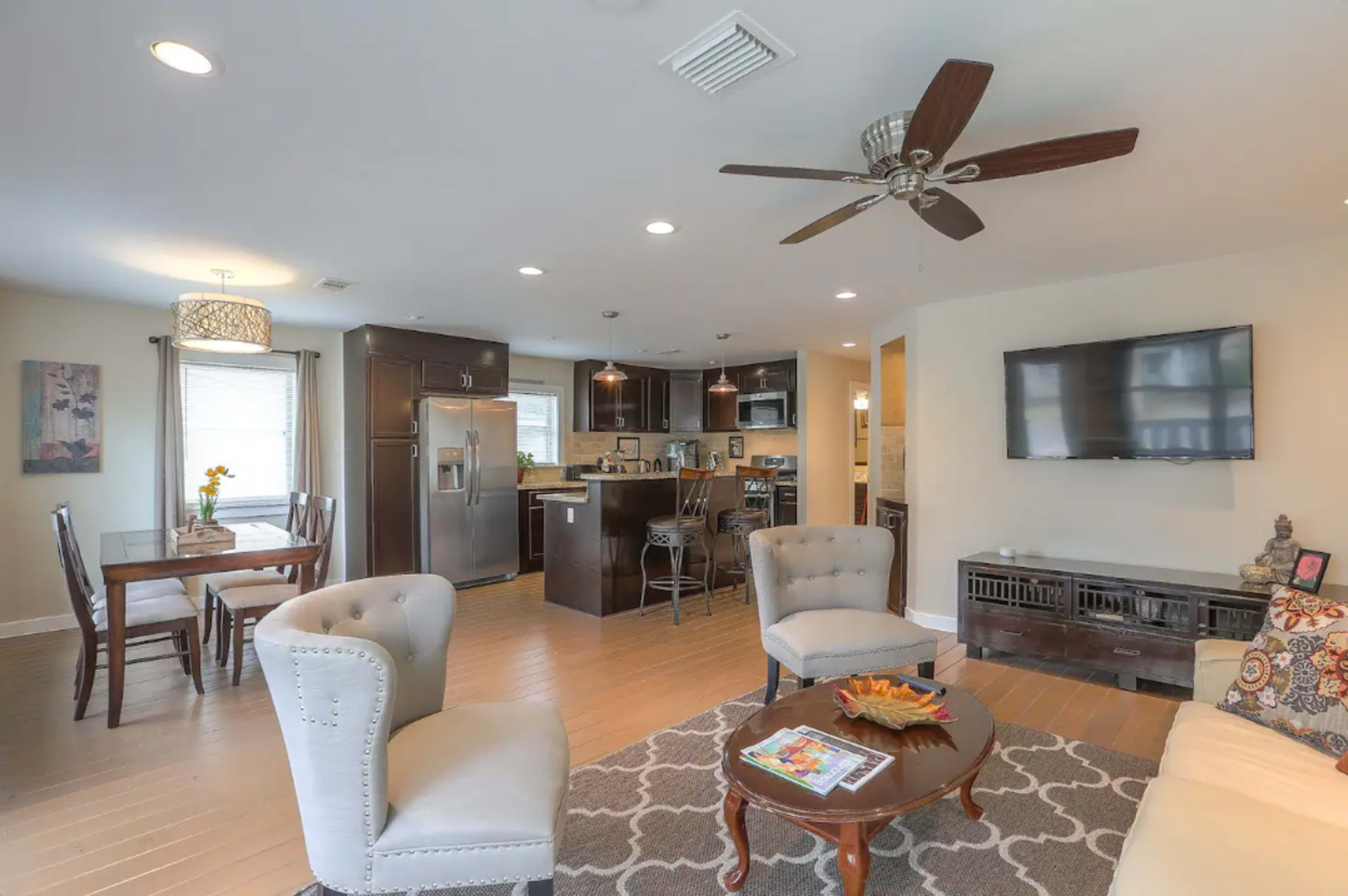 A modern open-concept living space featuring a kitchen, dining area, and seating area with a ceiling fan and a large flat-screen TV.