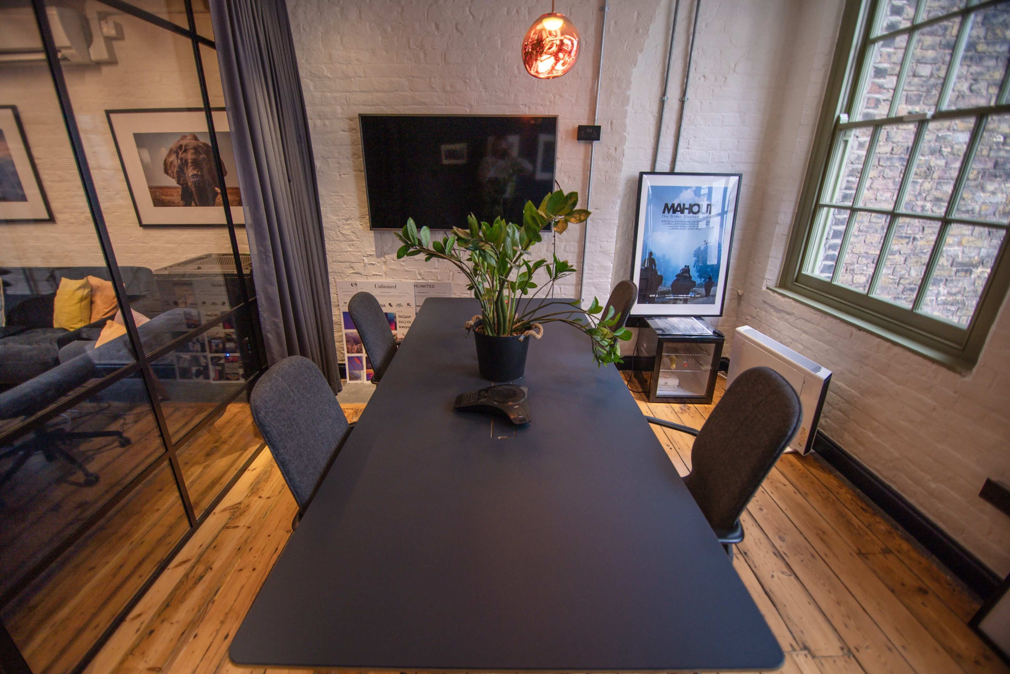 A modern conference room features a rectangular black table surrounded by four chairs, with a potted plant in the center and a wall-mounted television in the background.