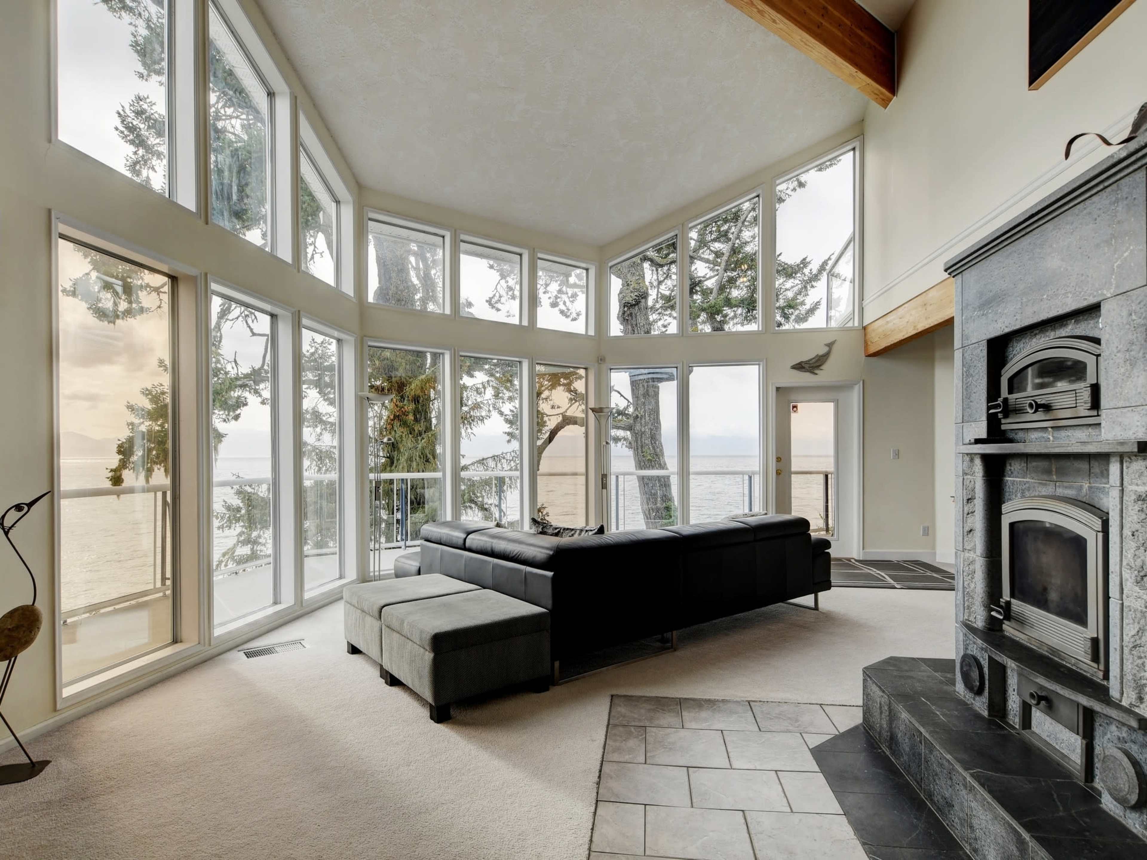 A modern living room features large windows overlooking a body of water, surrounded by trees, with a stone fireplace and a sectional sofa.