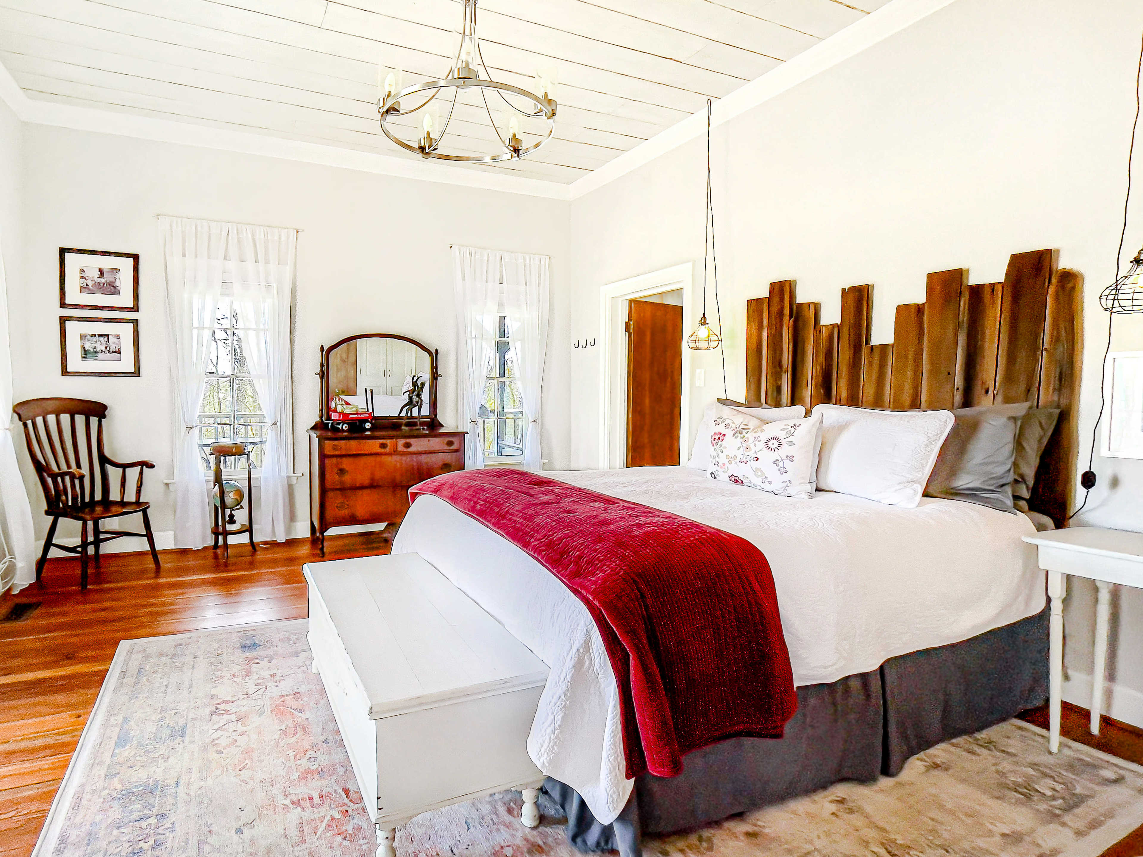 The image shows a bright, spacious bedroom featuring a large bed with a wooden headboard, a dresser, a rocking chair, and large windows with sheer curtains.