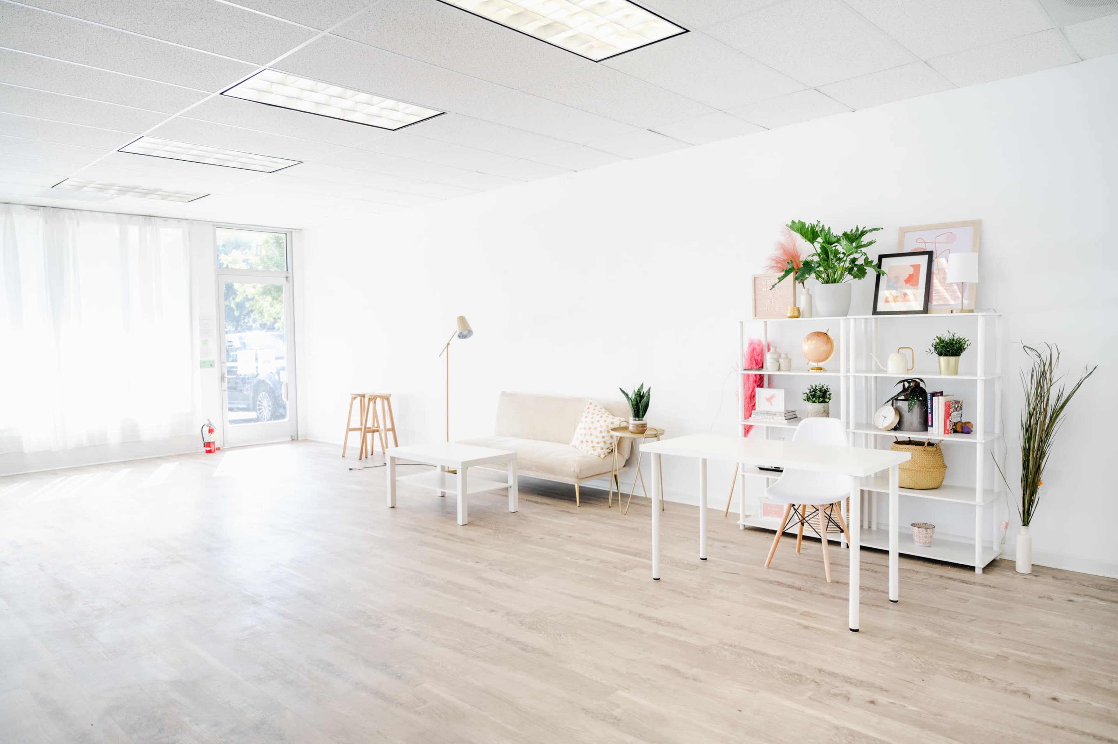 The image shows a bright, minimalistic room with light wooden flooring, featuring a beige sofa, a simple white bookshelf, and two white tables, along with natural light coming through large windows.