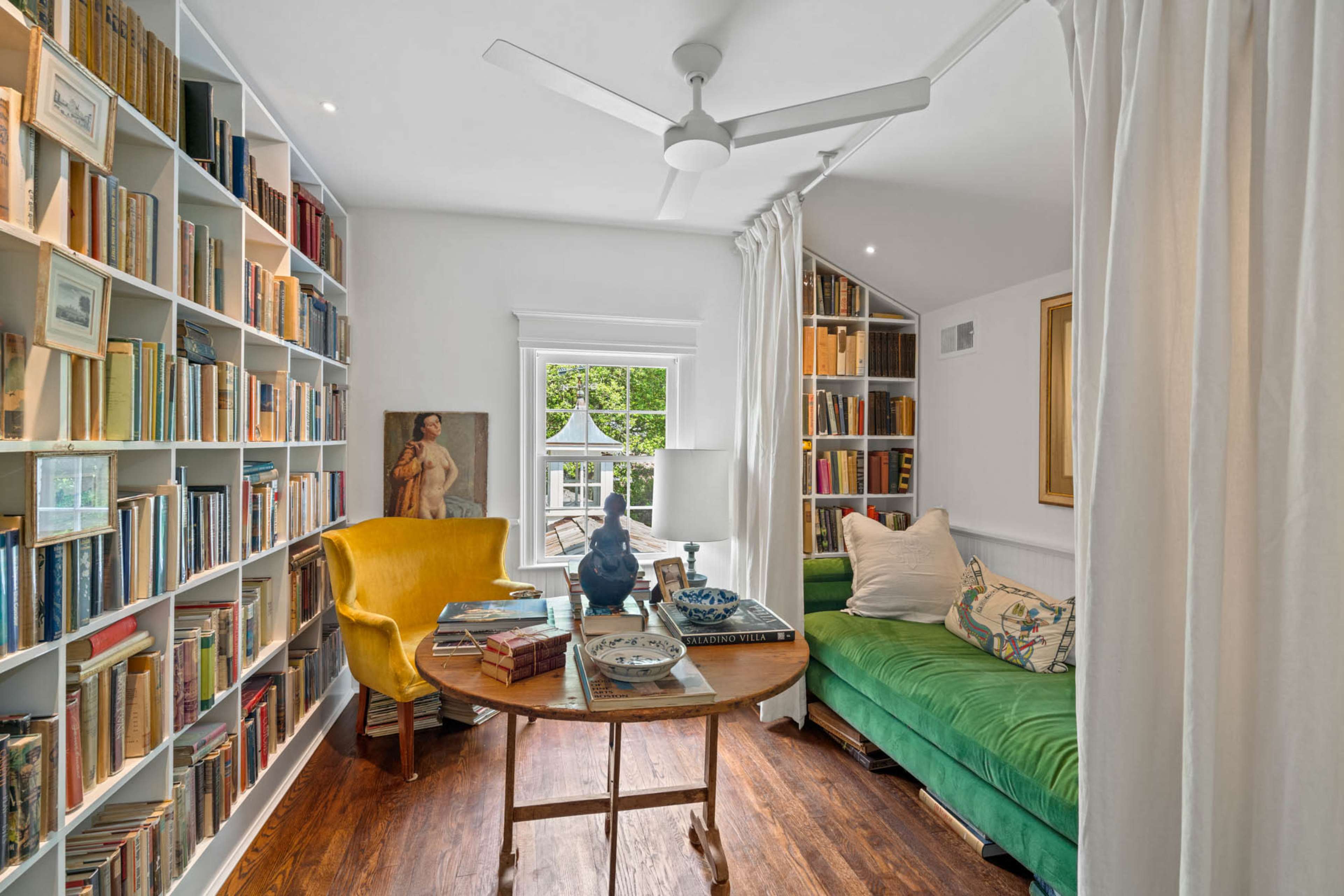 A cozy reading nook features a green sofa, a round table with books, and shelves filled with various books, all illuminated by natural light from a nearby window.