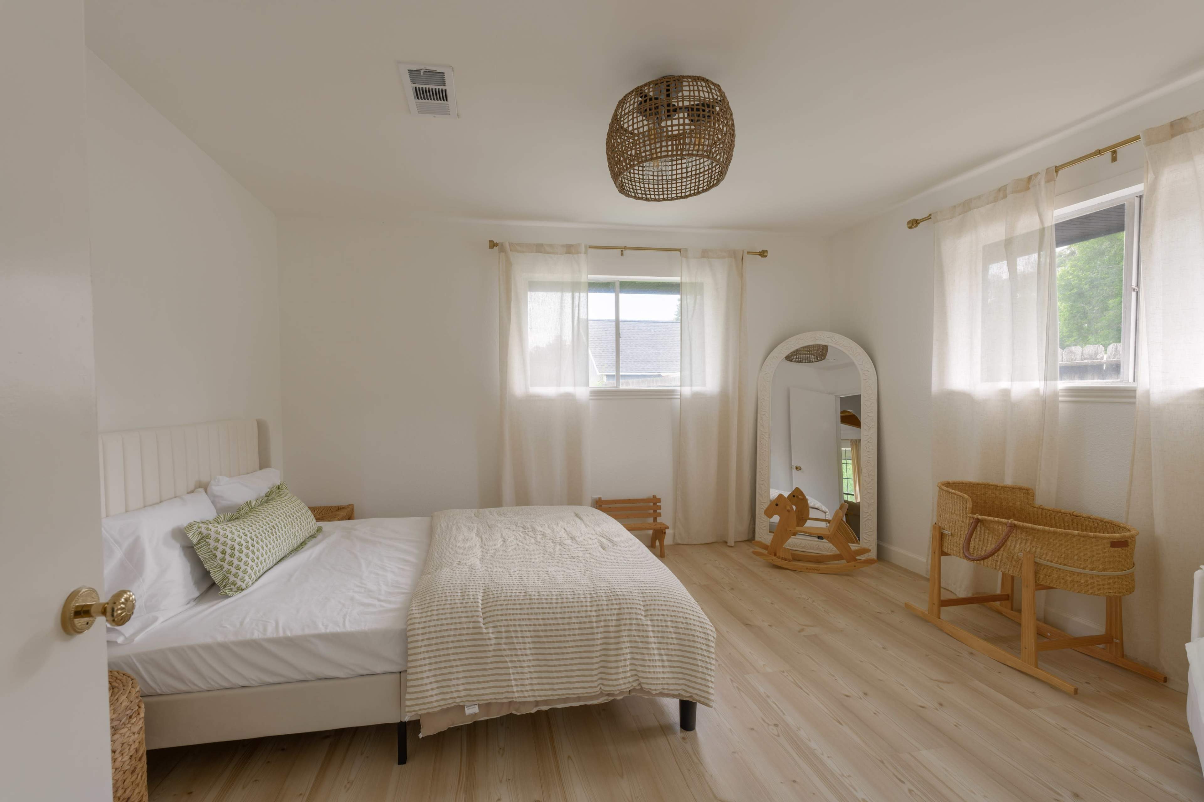 The image shows a minimalistic bedroom with a bed, a rocking horse, a mirror, and large windows covered with sheer curtains.