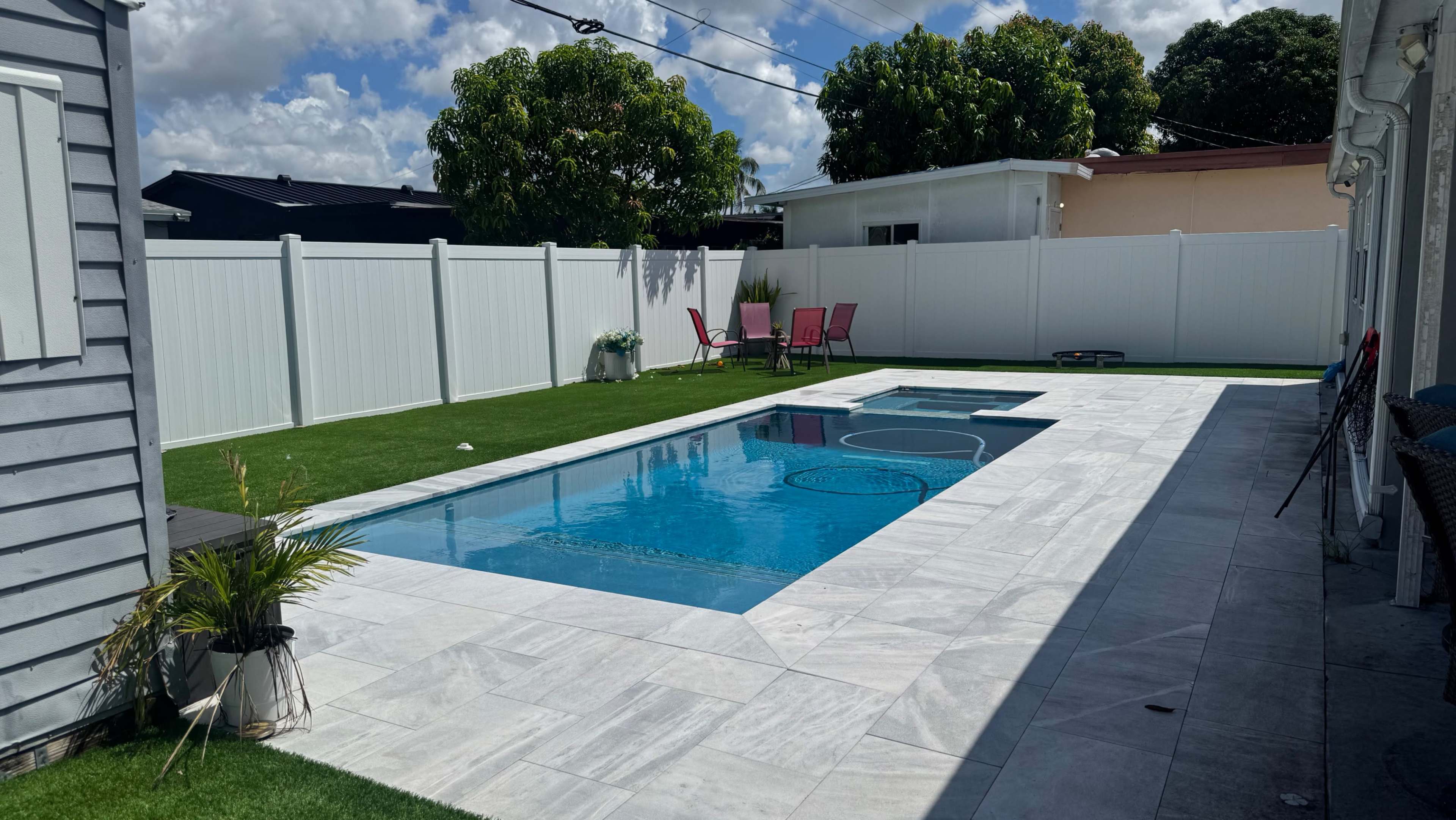 The image shows a rectangular swimming pool surrounded by a marble patio, grassy areas, and a white fence, with a few chairs visible nearby.