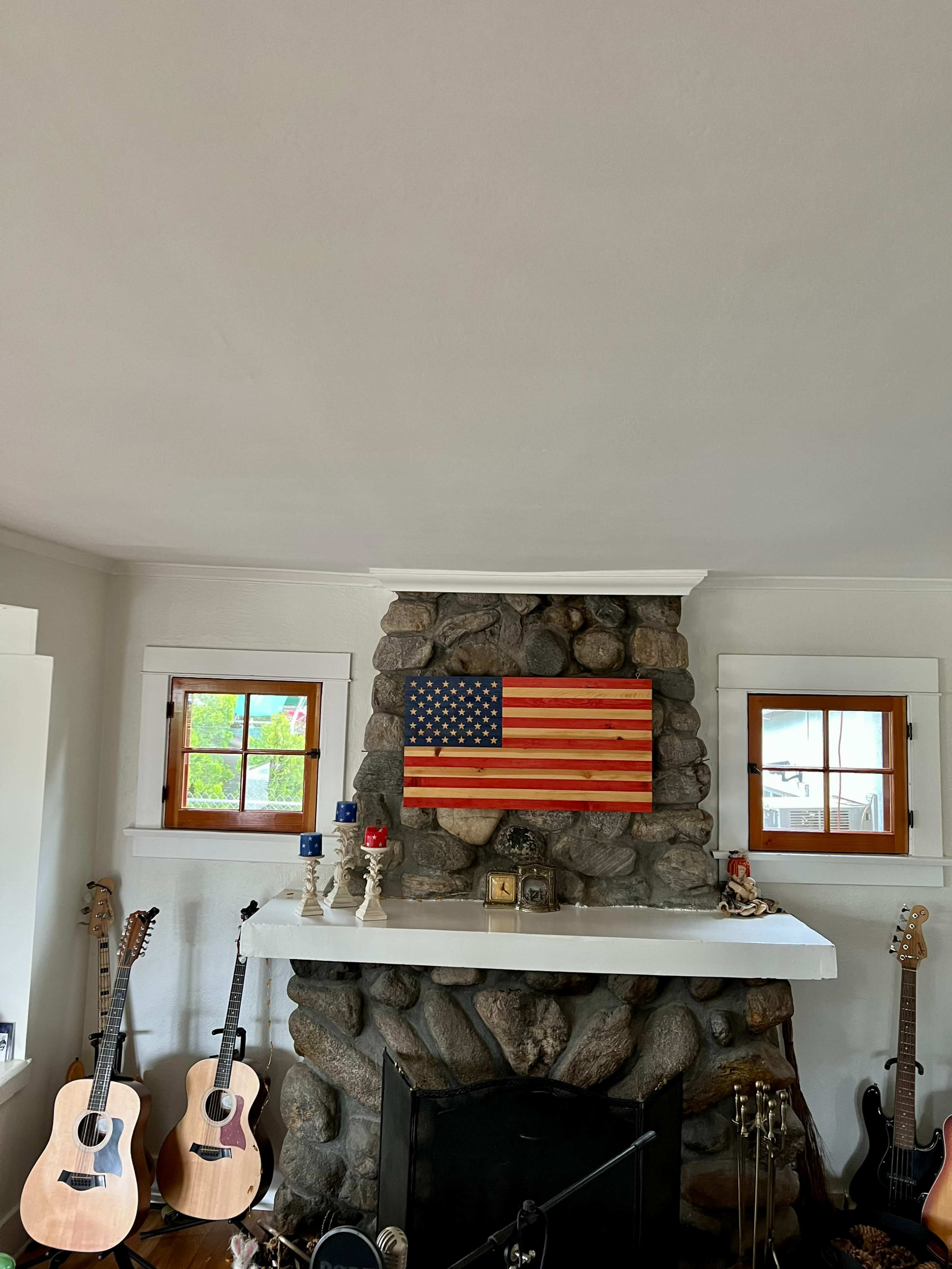 A stone fireplace is adorned with a wooden American flag, flanked by two windows and surrounded by acoustic guitars on either side.