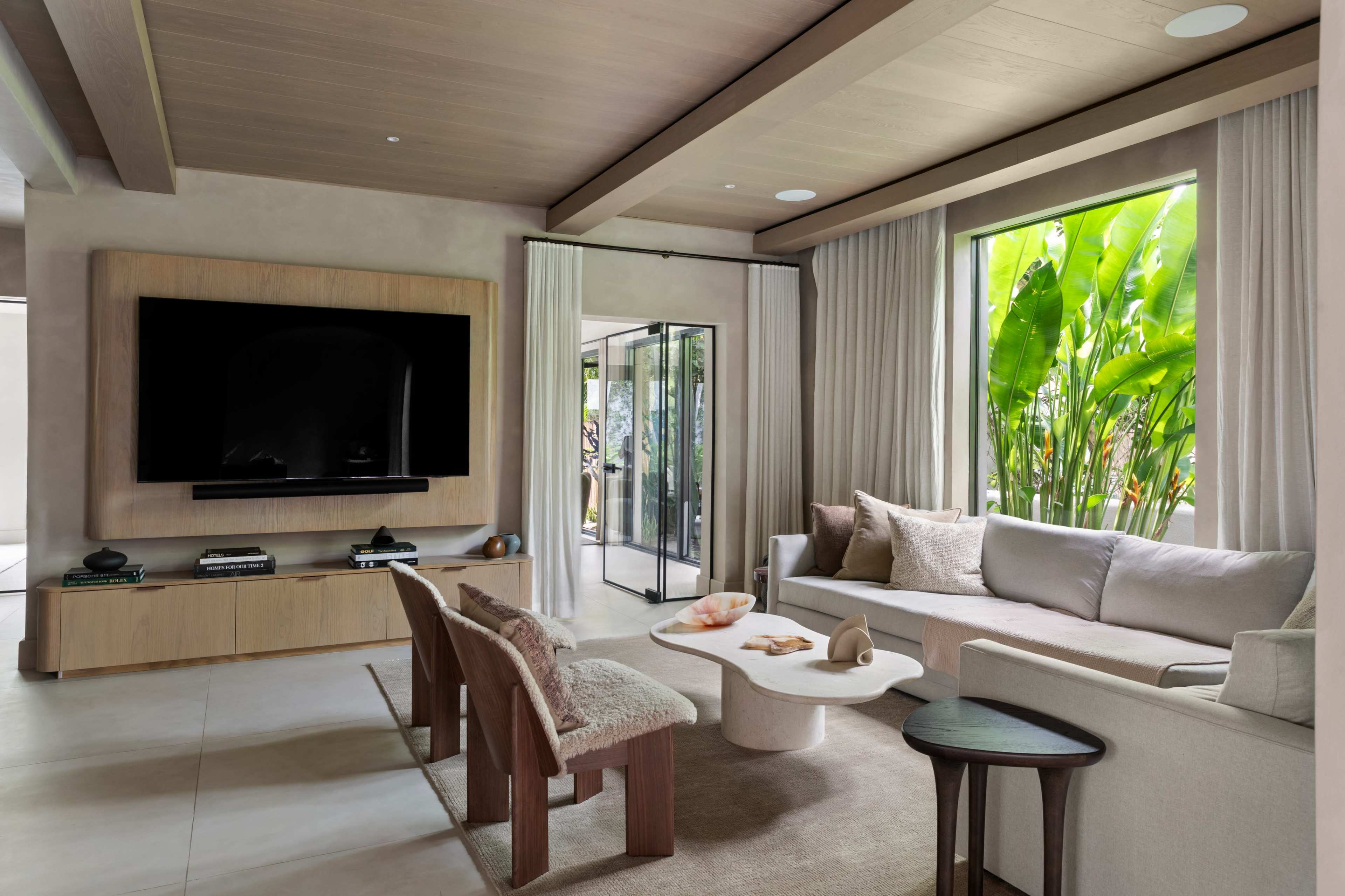 The living room features a modern design with a large television mounted on a wooden wall unit, a light-colored sectional sofa, and a round coffee table surrounded by two unique chairs, all set against a backdrop of lush greenery visible through large windows.