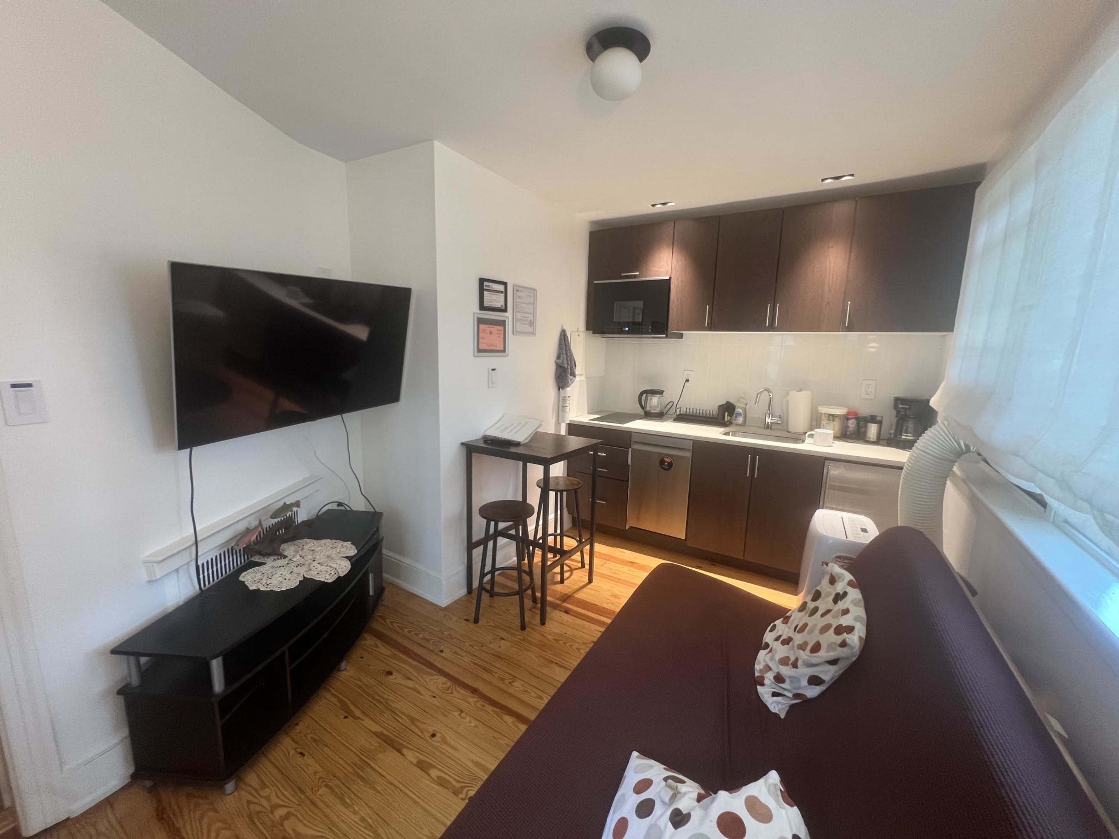 The image shows a compact kitchen and living area featuring a television, a small dining table, and modern appliances against a backdrop of wooden flooring.