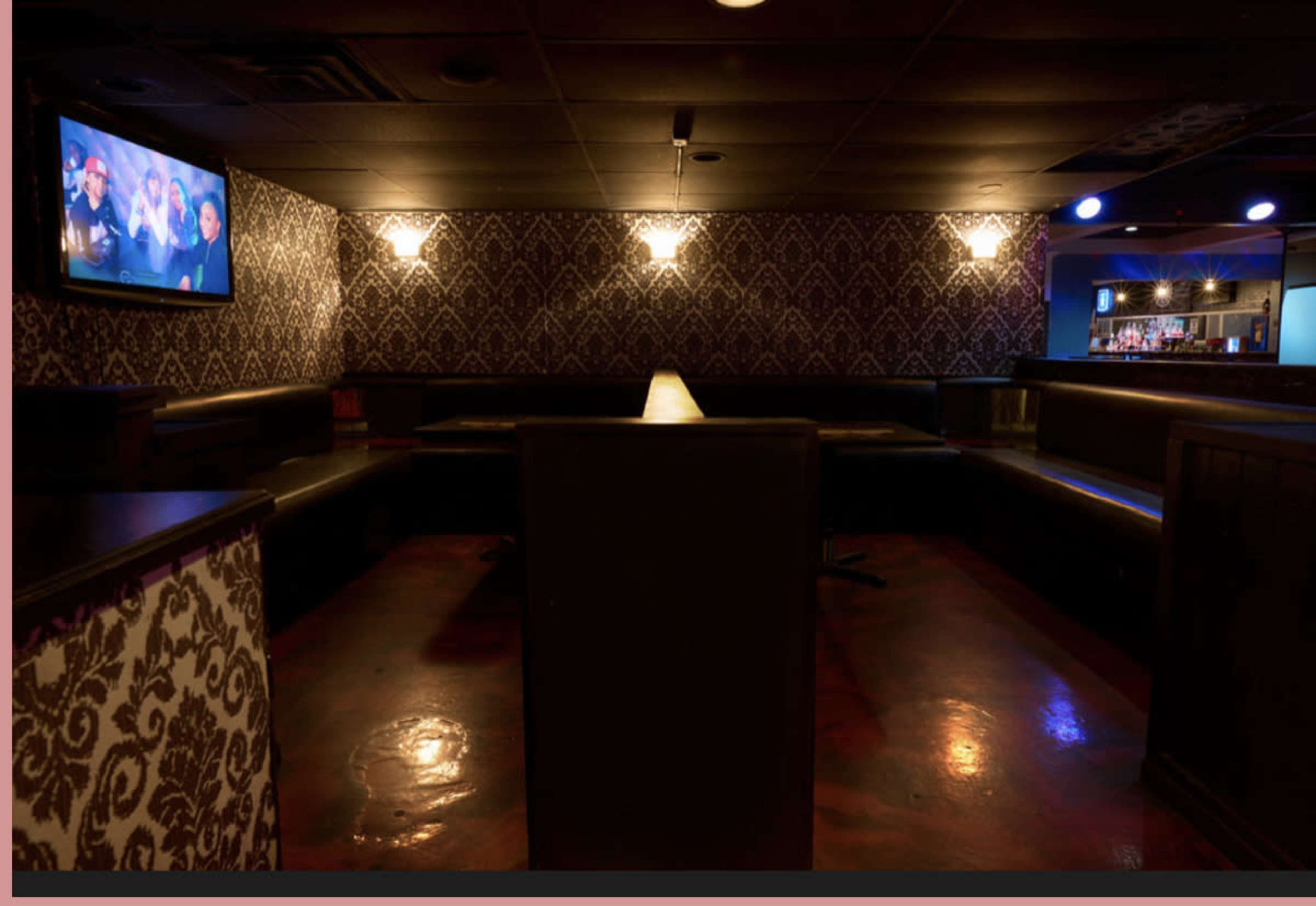 The image shows a dimly lit lounge area with black seating and a wall adorned with patterned wallpaper, featuring a television on one side.