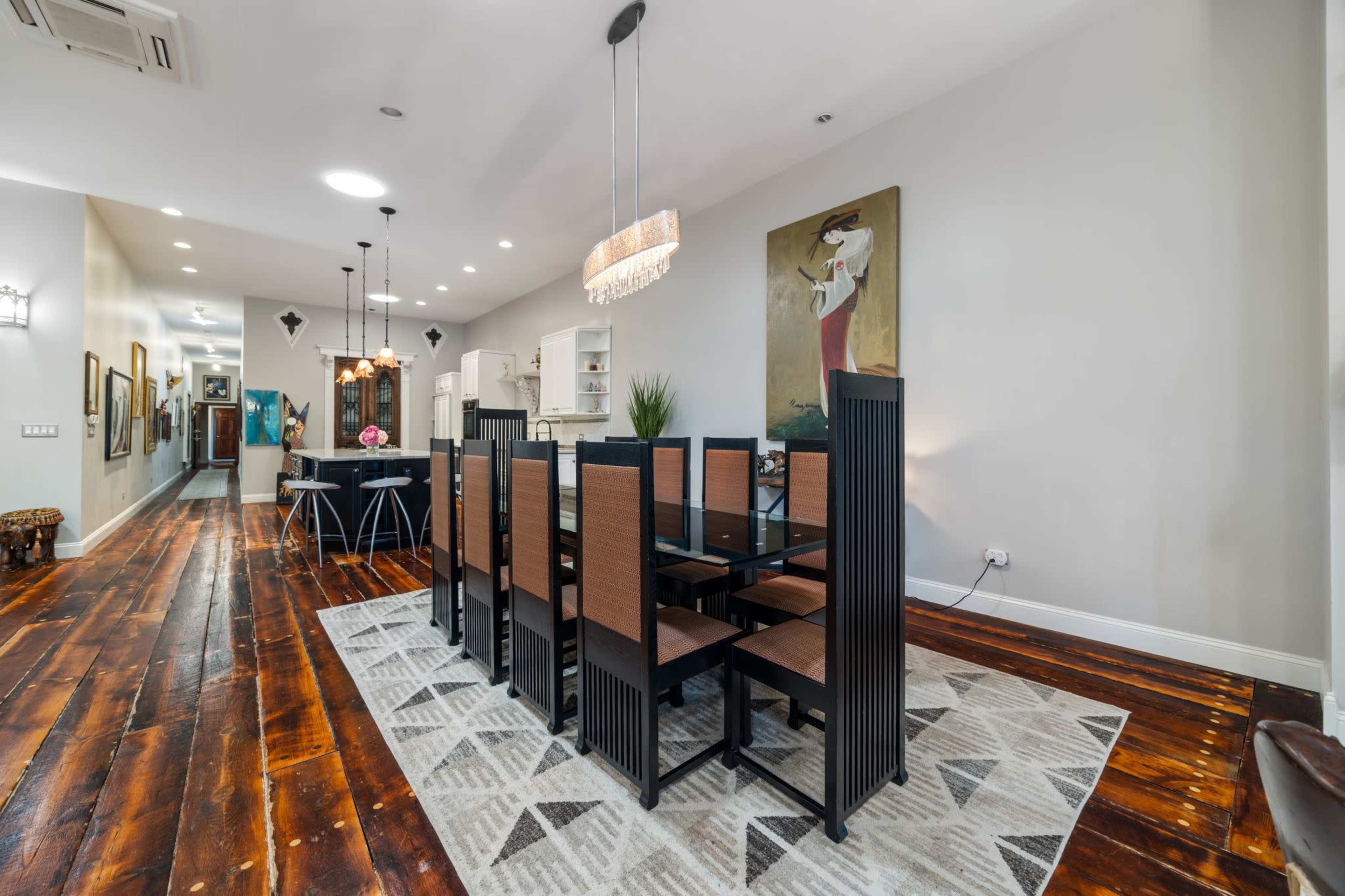 The image shows a modern dining area with a long table surrounded by eight chairs, set against a backdrop of hardwood flooring and decorative artwork on the walls.