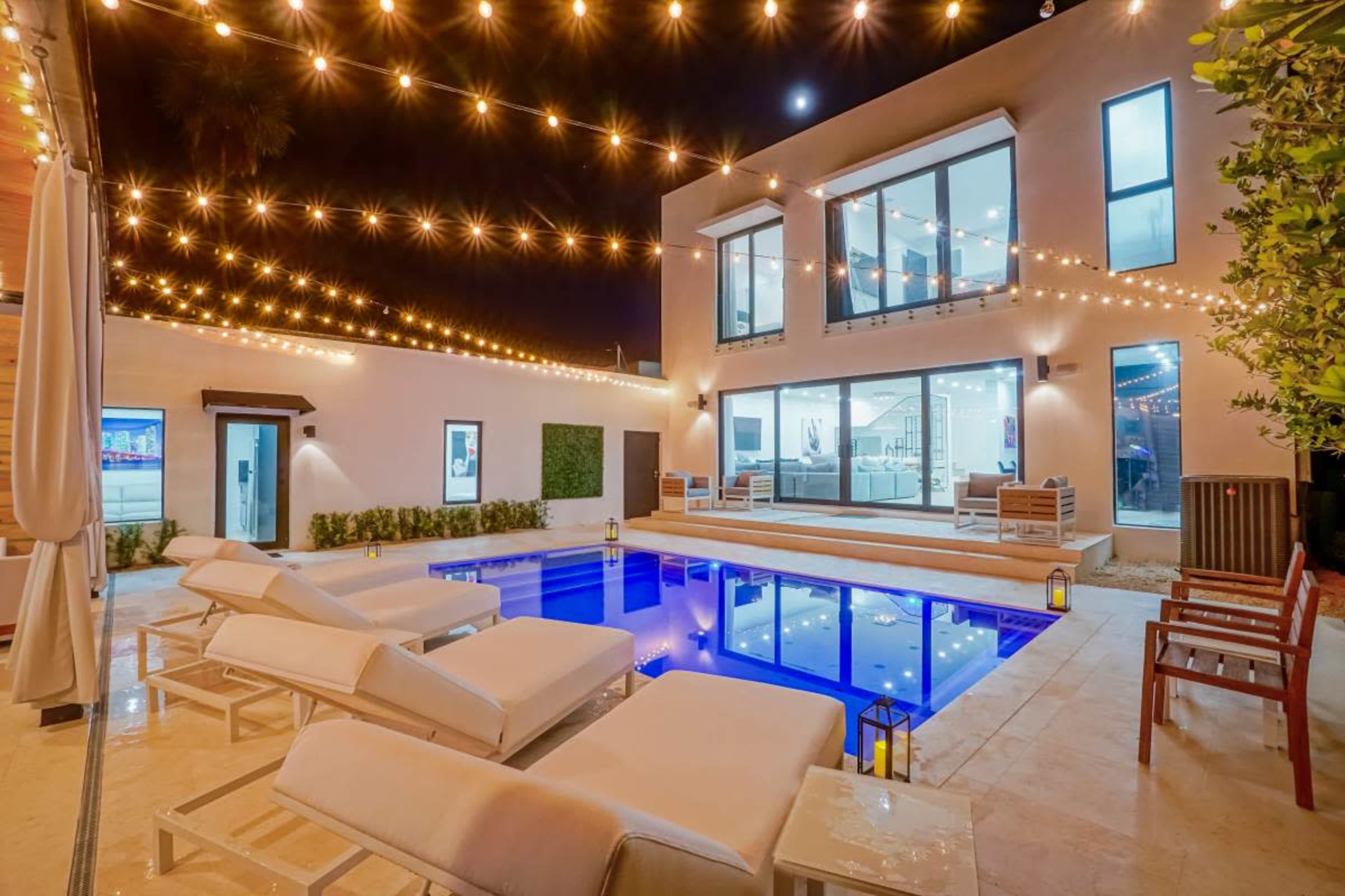 A modern backyard features a pool illuminated by string lights, surrounded by lounge chairs and a view of a two-story house.