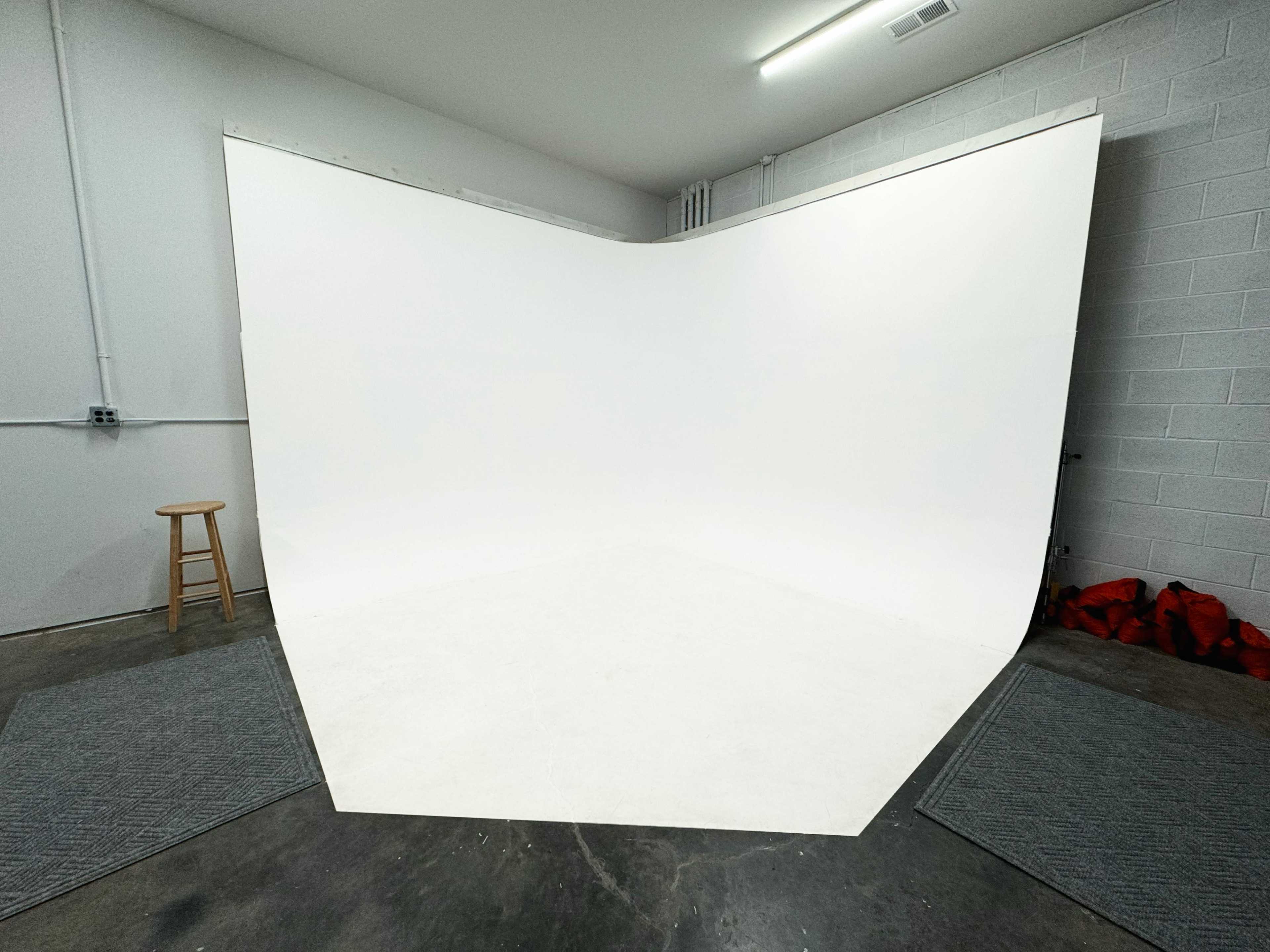 A spacious studio featuring a large seamless white backdrop, gray mats, and a wooden stool positioned in the corner.