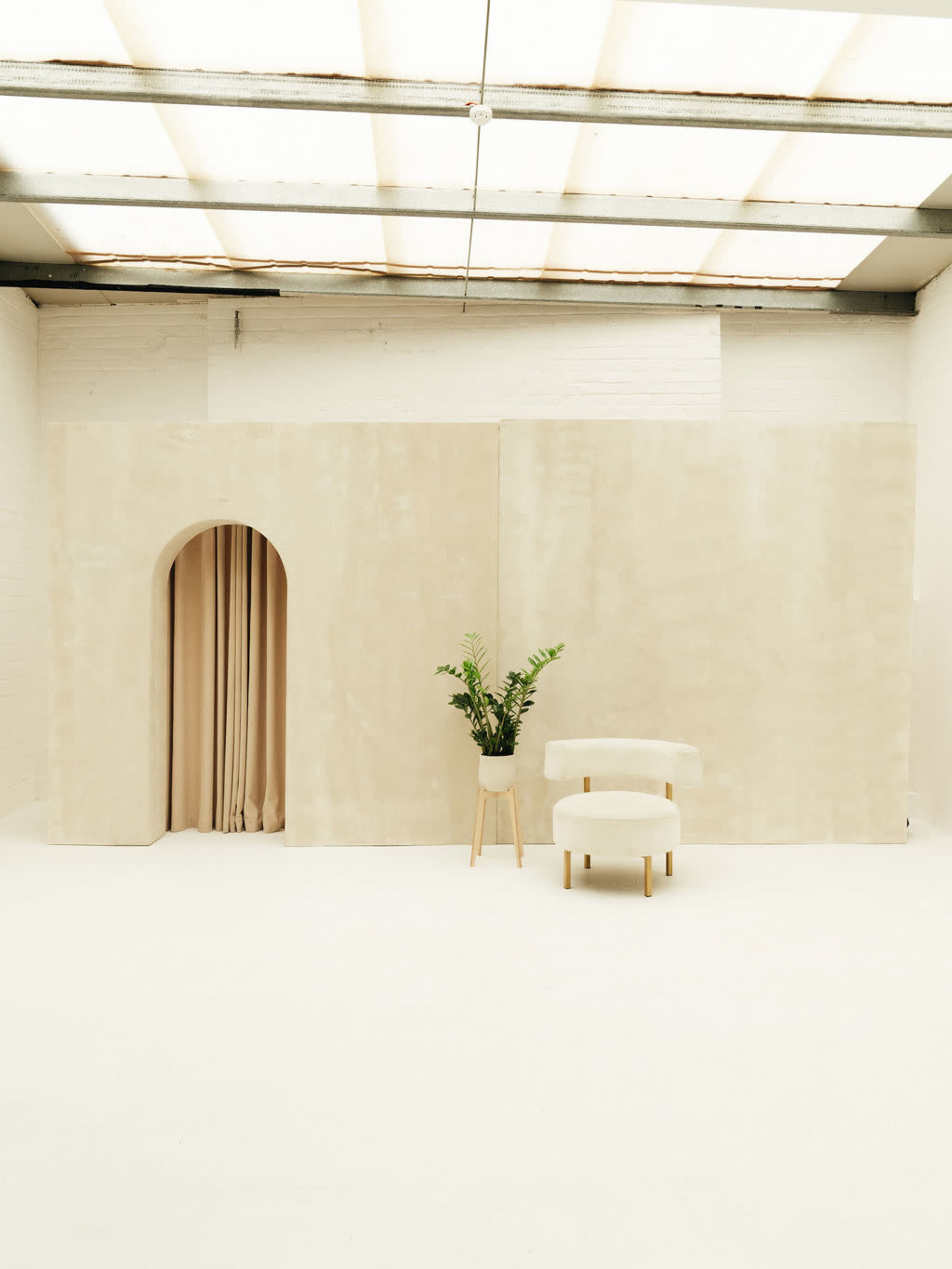 The image shows a minimalistic interior space with a beige wall, an archway leading to draped fabric, a potted plant, and a simple chair.