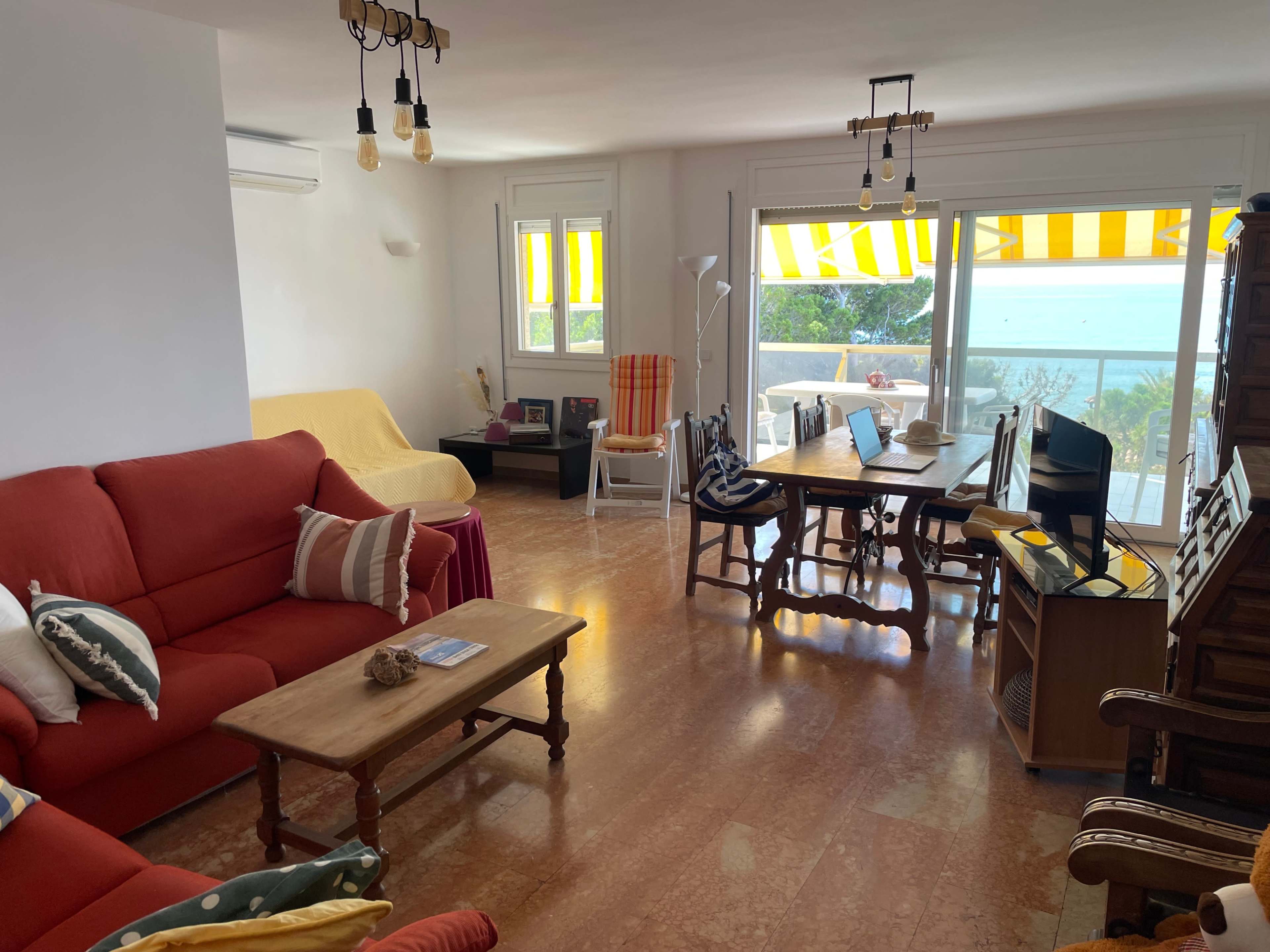 The room features a red sofa, a wooden dining table with chairs, and large windows showcasing a view of the sea, with yellow striped awnings filtering the light.